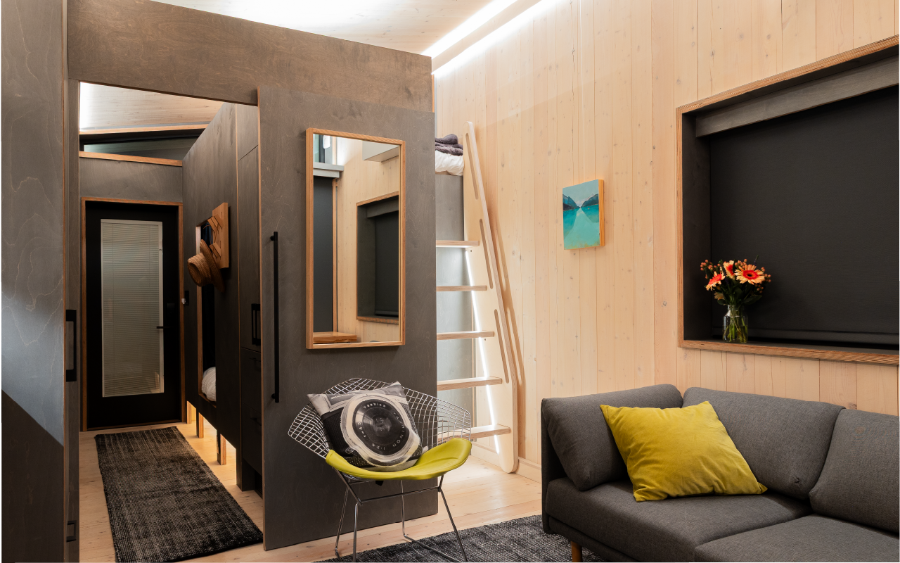 Interior of a modern tiny house with a gray couch, a yellow pillow, a small shelf with a vase of flowers, and a ladder leading to a lofted bed. There is a door, a mirror, and a decorative wall shot