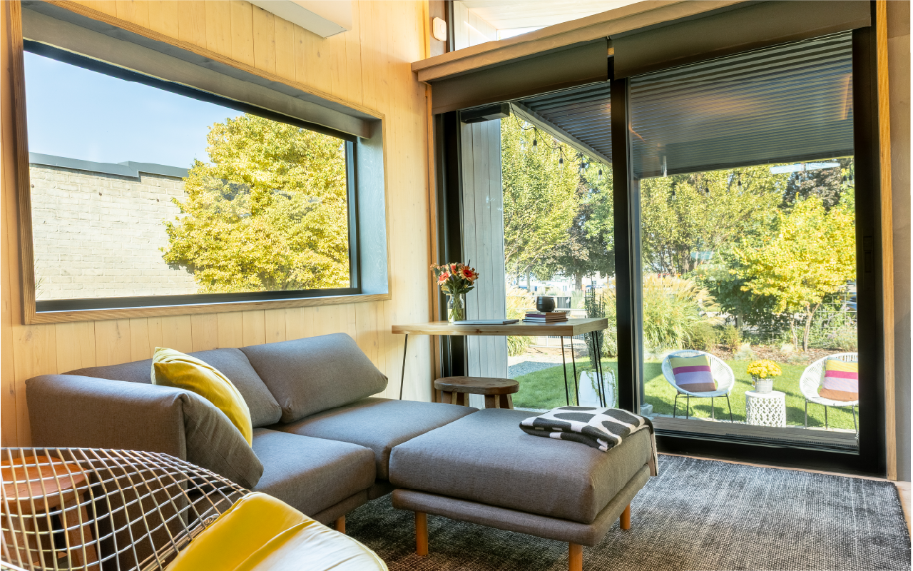 Modern living room with large windows overlooking a garden with trees and outdoor chairs, featuring a gray sectional sofa with yellow pillows and a throw blanket, a small round wooden table with a vase of flowers, and a glass sliding door leading to 