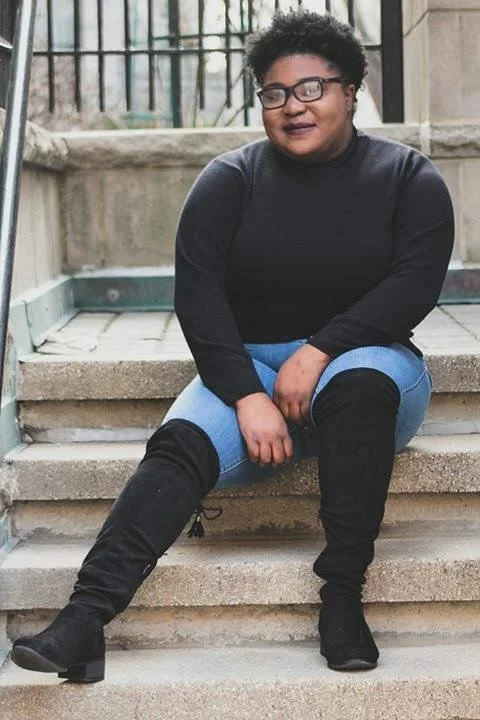 A person sitting on concrete steps outdoors, wearing glasses, a black long-sleeve shirt, blue jeans, and knee-high black boots.