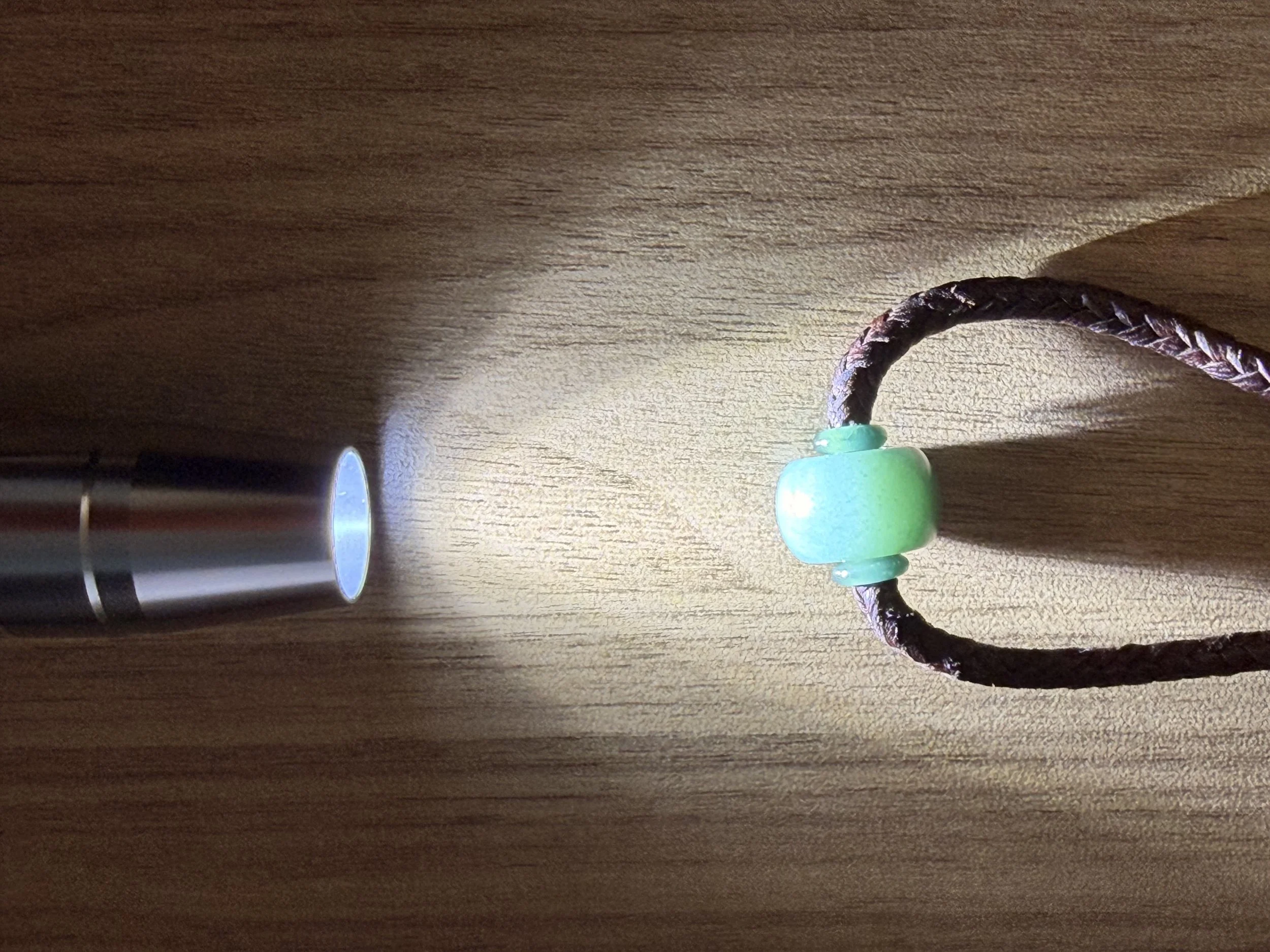 A flashlight shining a beam of light onto a choker necklace with a jadeite jade bead, resting on a wooden surface.