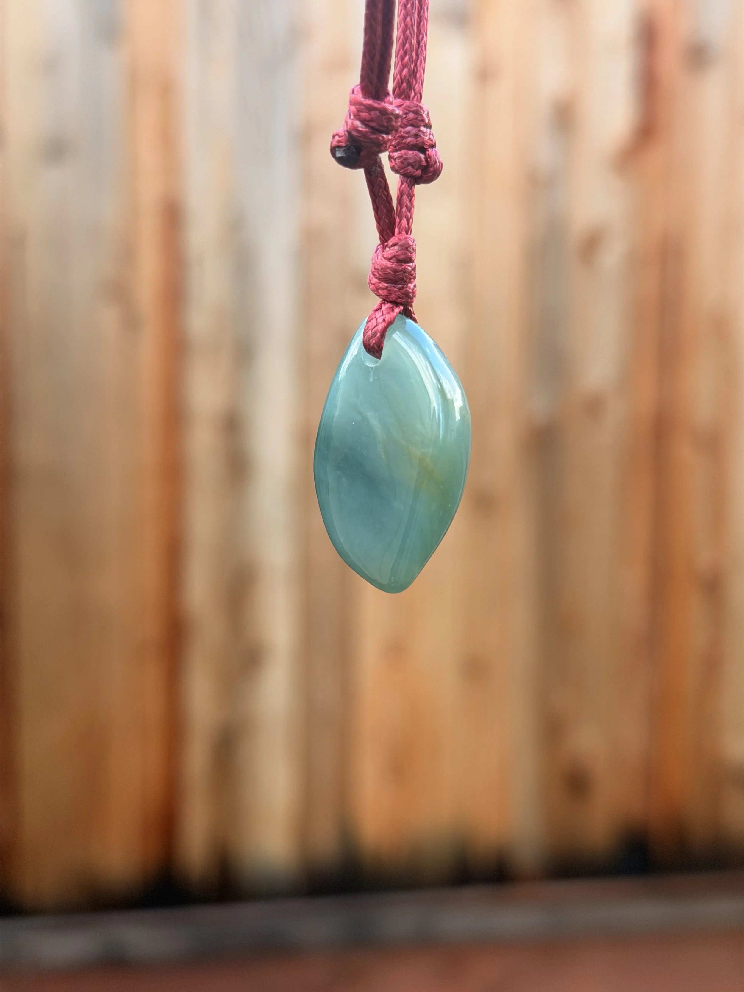 A teardrop-shaped, polished greenish-blue jadeite jade pendant hanging from a pink cord with knots, against a blurred wooden background.