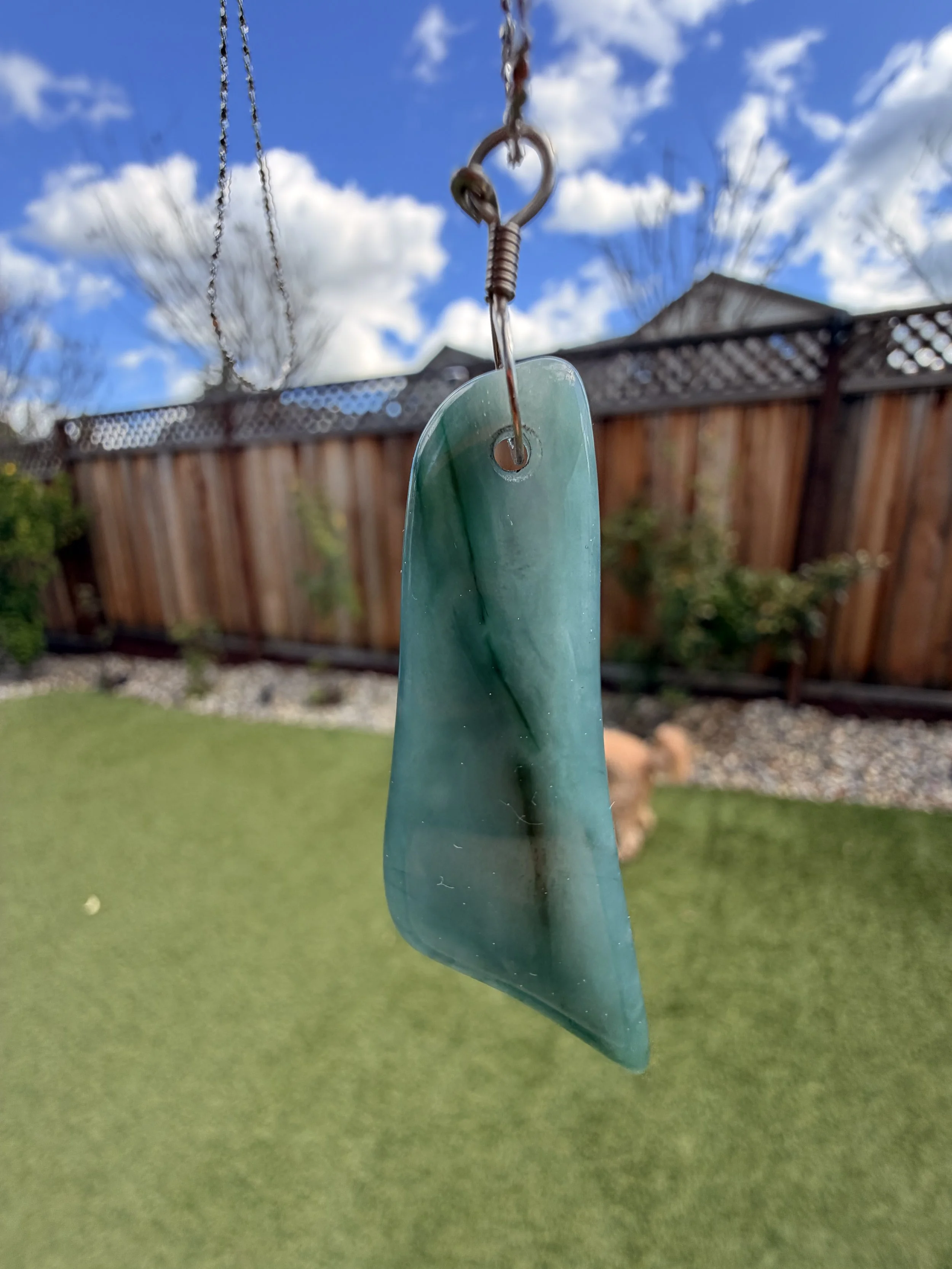 A close-up of a sail or wind chime made from a piece of sea glass hanging from a chain outdoor with a wooden fence, green grass, and a blue sky with clouds in the background.