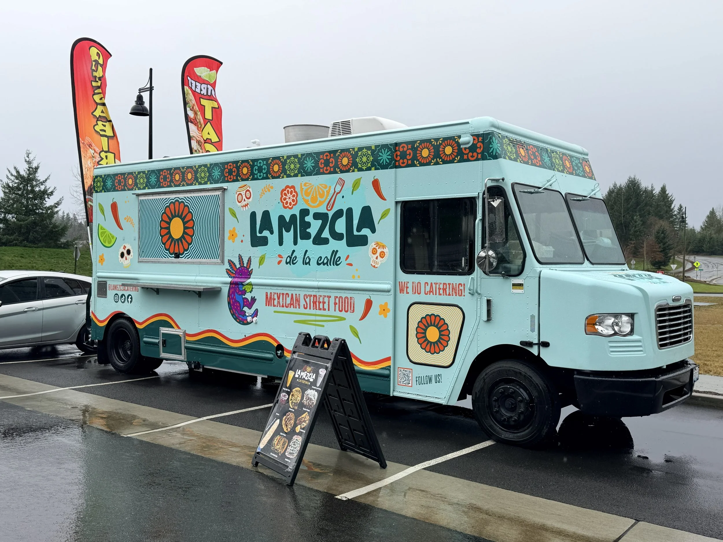 Colorful Mexican food truck with signage and flags in front of a parking lot, on a rainy day.