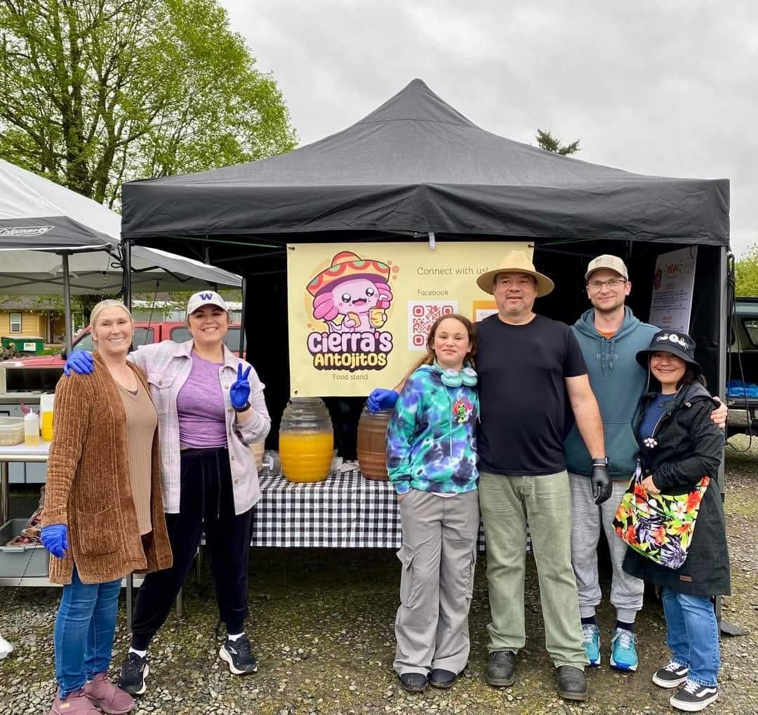 Group of six people standing in front of a food stand at an outdoor event, with a black canopy tent and a yellow sign that reads 'Cierra's Antojitos,' a cartoon character mascot, and QR codes for social media. The people are smiling, some wearing gloves and aprons, indicating they might be vendors or staff.