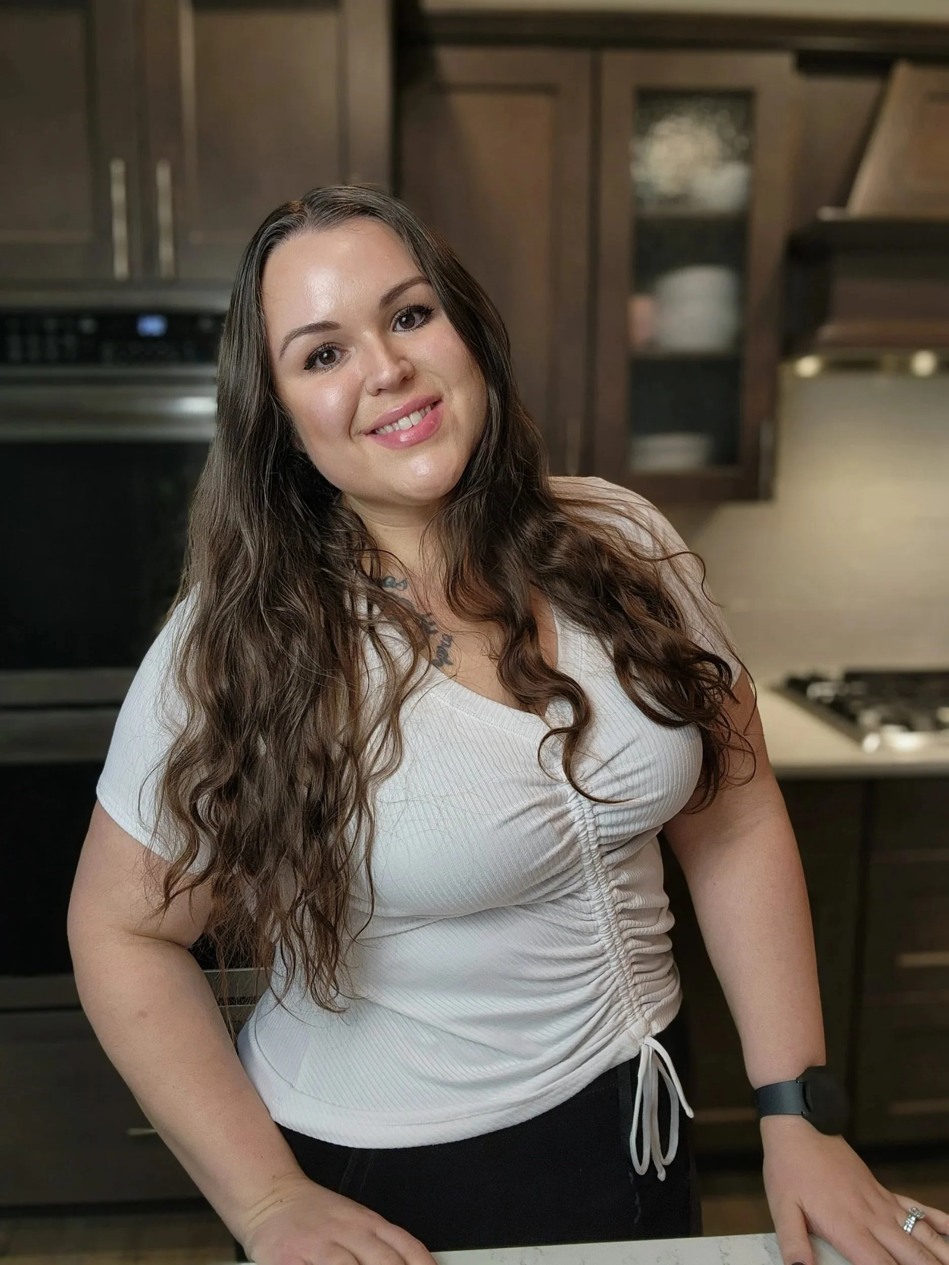 Woman with long, wavy brown hair wearing a white top and black pants, standing in a kitchen.