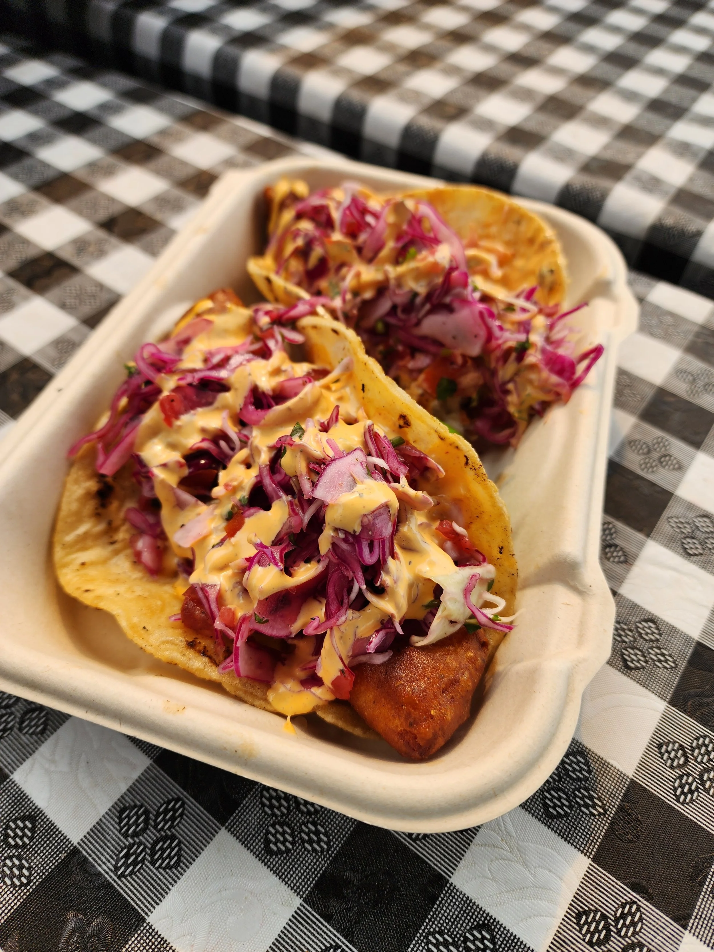 Close-up of two fish tacos topped with shredded red and green cabbage and drizzled with sauce, in a disposable container on a checkered tablecloth.