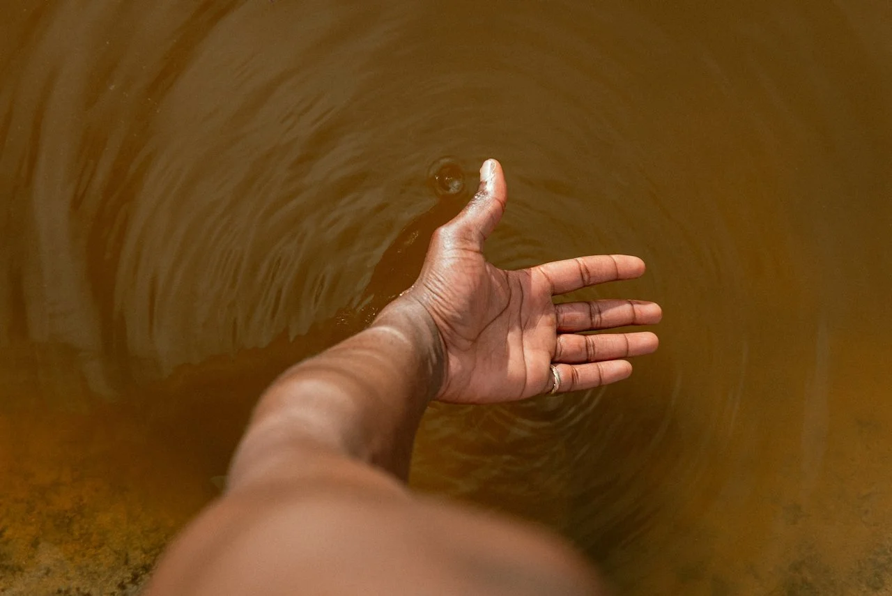 A person's hand reaching into a brown body of water, creating ripples around the hand.