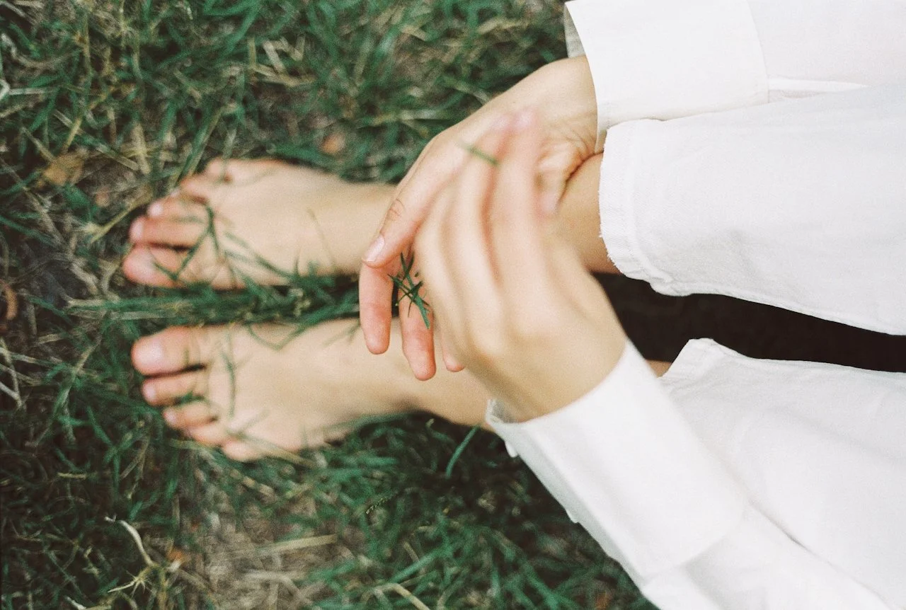 Person sitting on grass with feet visible, wearing a white long-sleeve shirt, holding a small green plant or grass between their fingers.