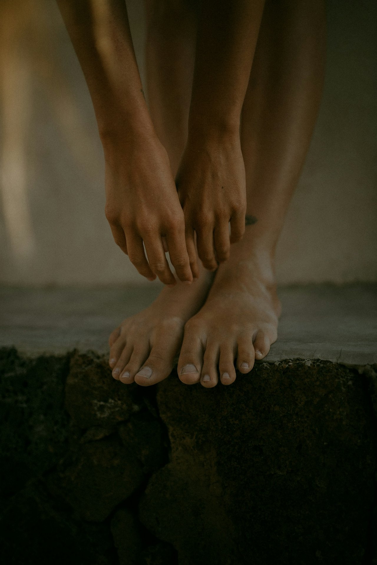 Close-up of hands and feet, with one person standing on a dark stone surface while holding their ankles, in a minimalistic or artistic setting.