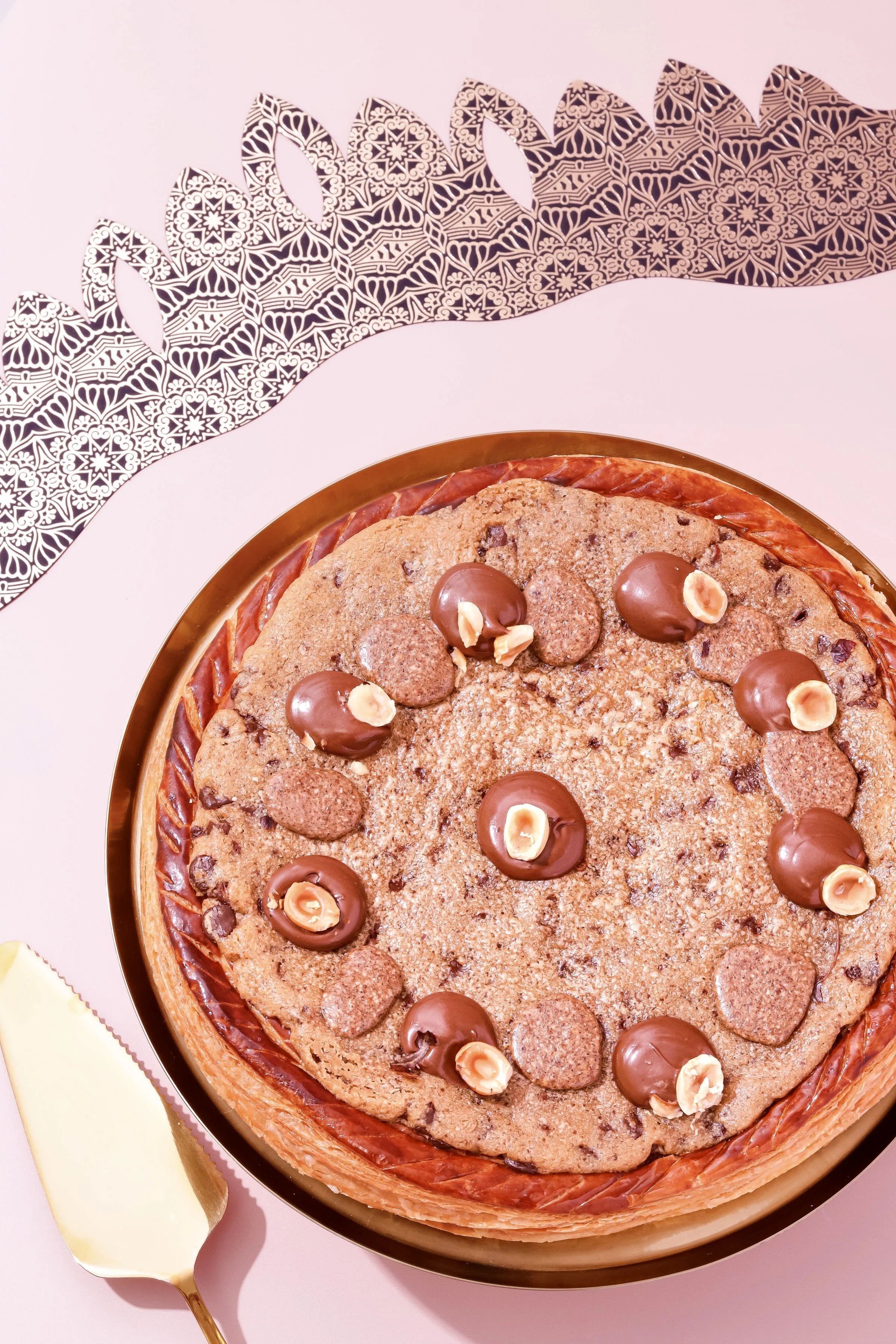 Gâteau aux pépites de chocolat décoré de chocolats et de noisettes, présenté dans un plat rond avec un fond rose clair et une décoration en papier à motif.