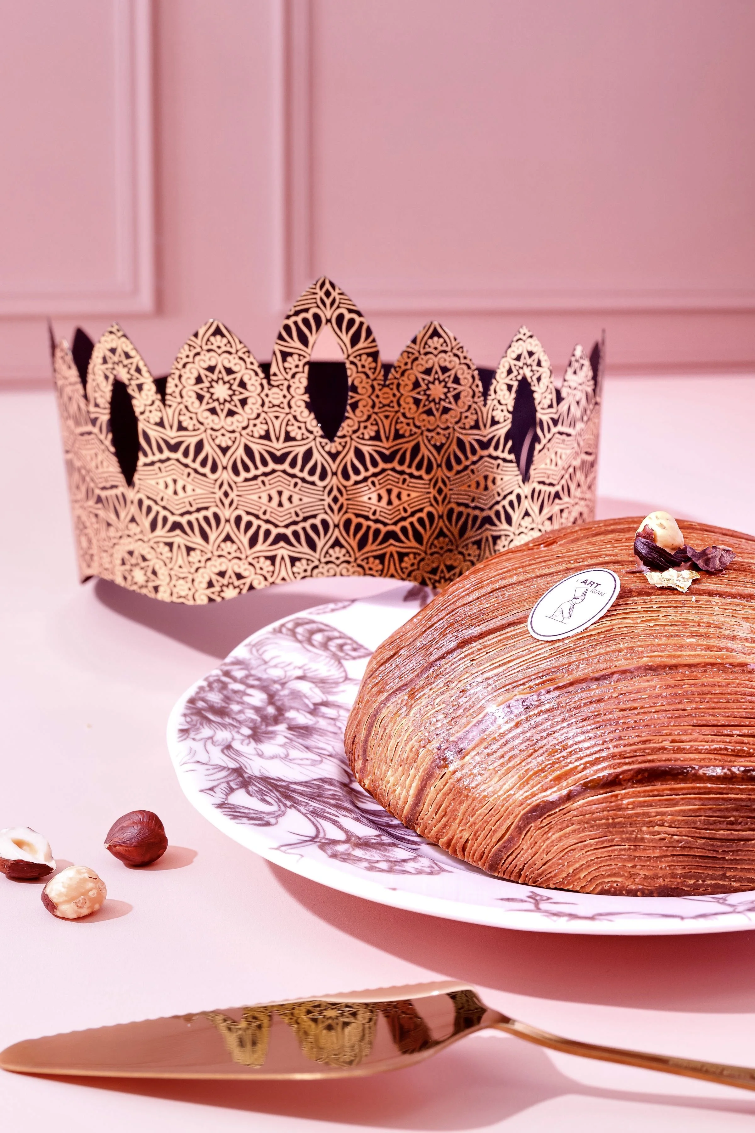 Gâteau en forme de coque avec une décoration de petits morceaux de chocolat et noisettes, placé sur une assiette blanche avec un motif d'arbre, et une couronne en papier métallique dorée en arrière-plan, sur une table rose avec une cuillère dorée à côté.