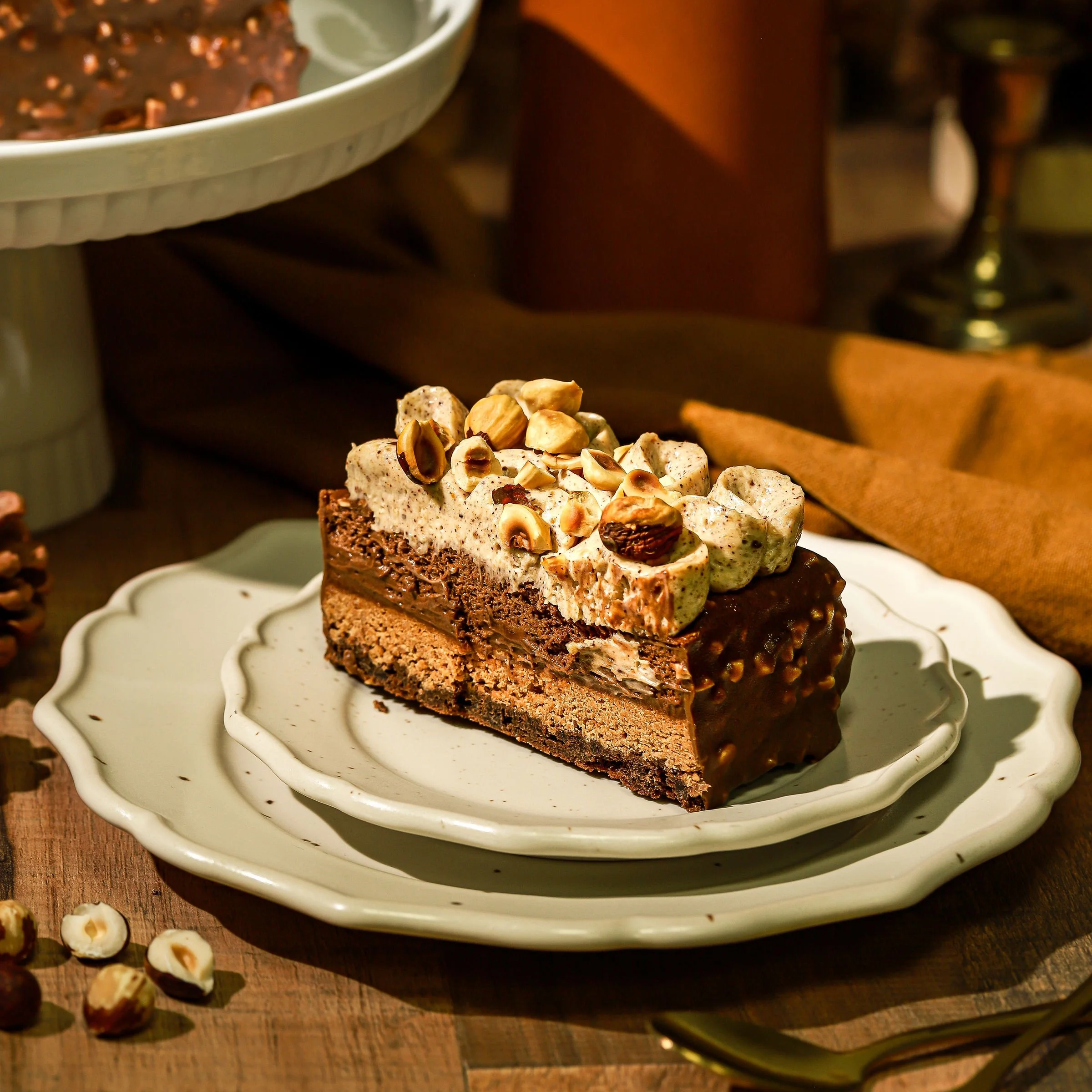 Une part de gâteau au chocolat avec des noisettes sur le dessus, sur une assiette blanche suvère, avec des noisettes autour. En arrière-plan, un plateau avec plus de gâteau et un tissu marron.