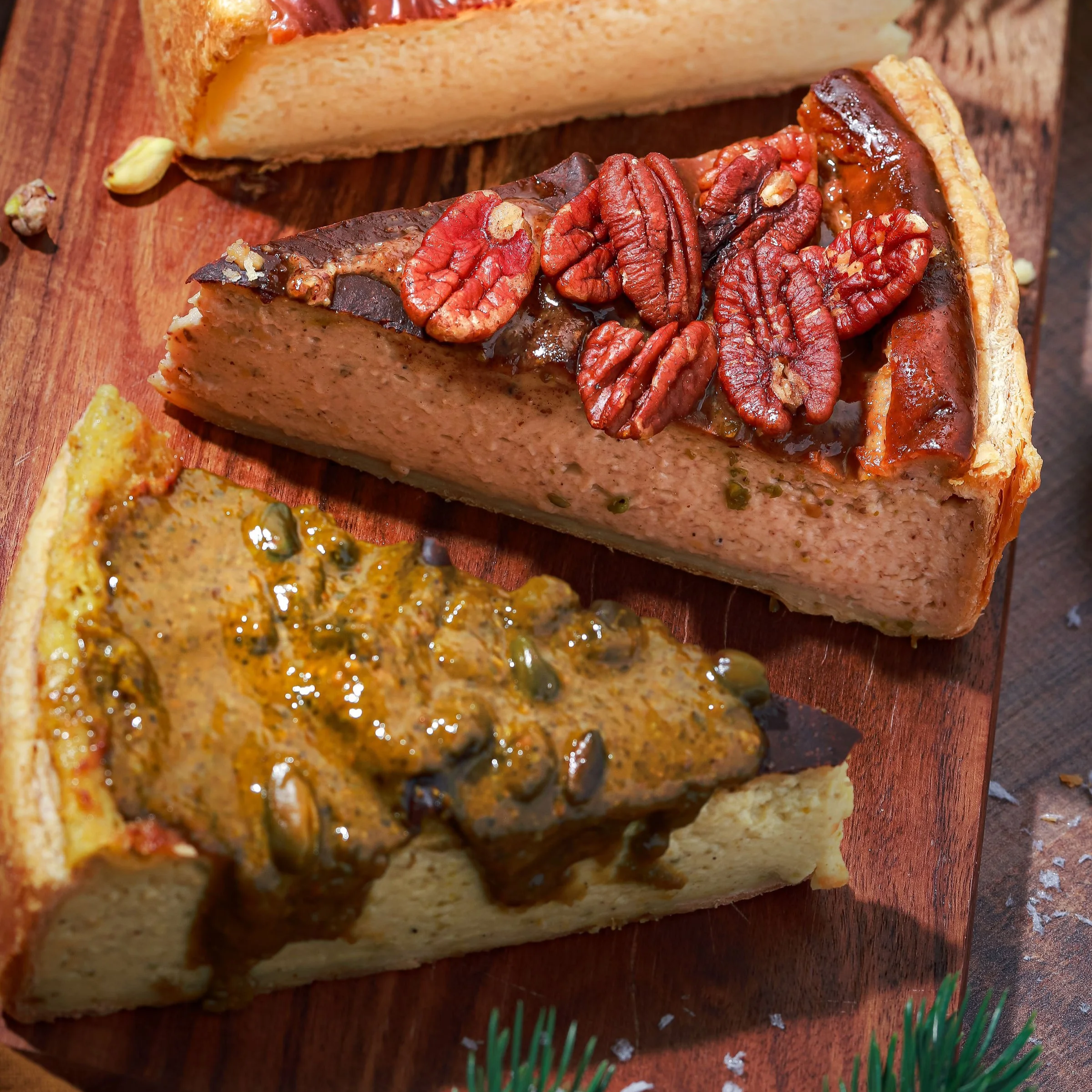 Trois tranches de terrine avec différentes garnitures sur une planche en bois, garnie de noix, de sauce et de graines.