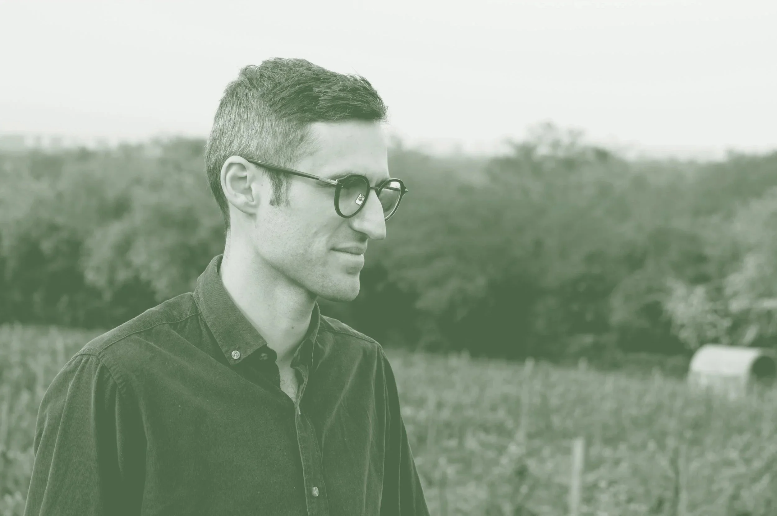 Jeune homme avec lunettes, versant de l'eau dans sa bouche, en extérieur dans un champ avec un arbre en arrière-plan.