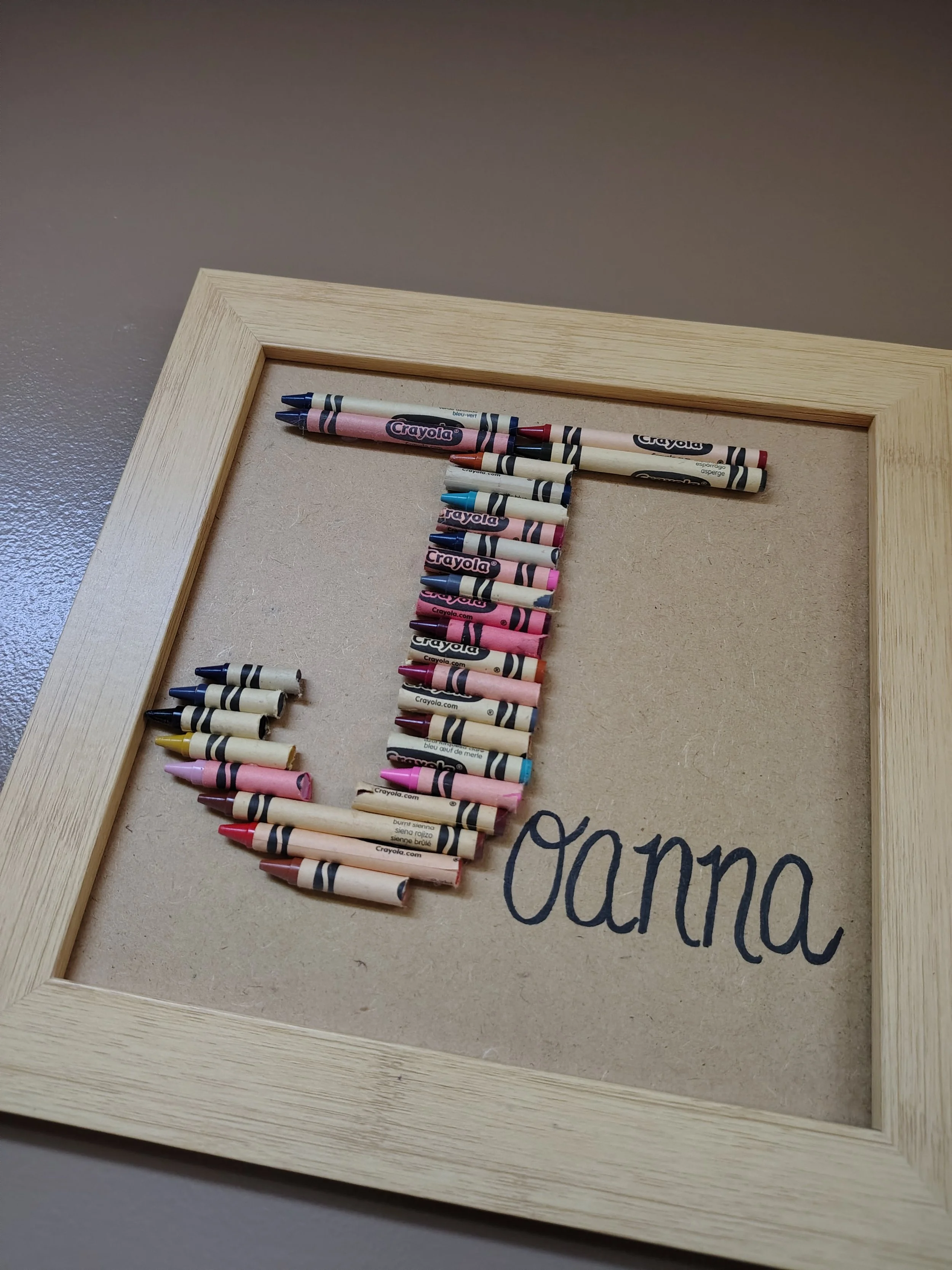 A corkboard framed with light wood, arranged to form the letter 'J' using crayon pieces, with the word 'Oanna' handwritten in black ink beneath.