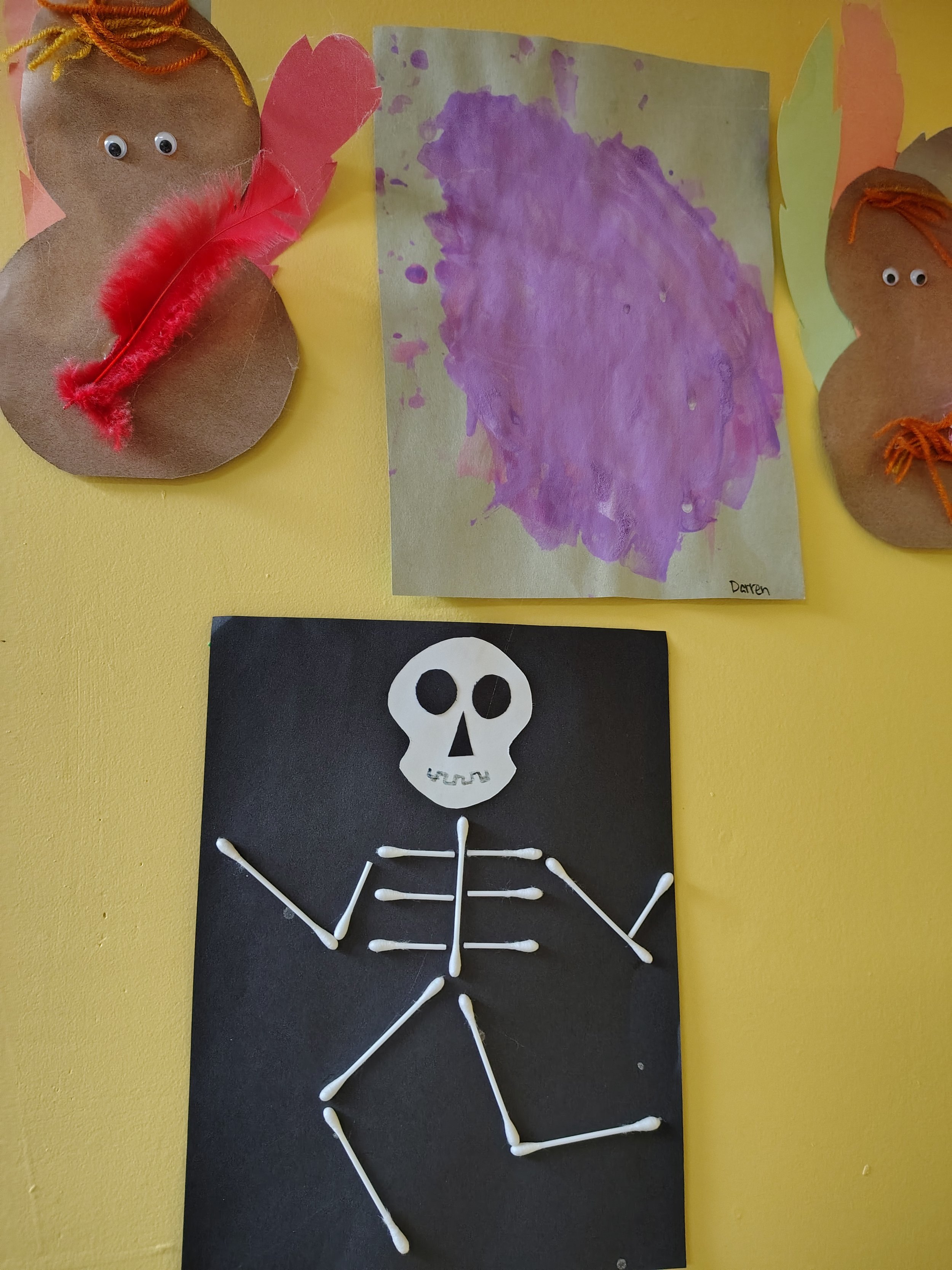 Child's artwork on a yellow wall: a purple watercolor painting of a large, amorphous shape labeled 'Darren'; a black poster with a white skeleton made of cotton swabs; two brown, paper-mache snowman crafts with googly eyes, orange yarn hair, and pink