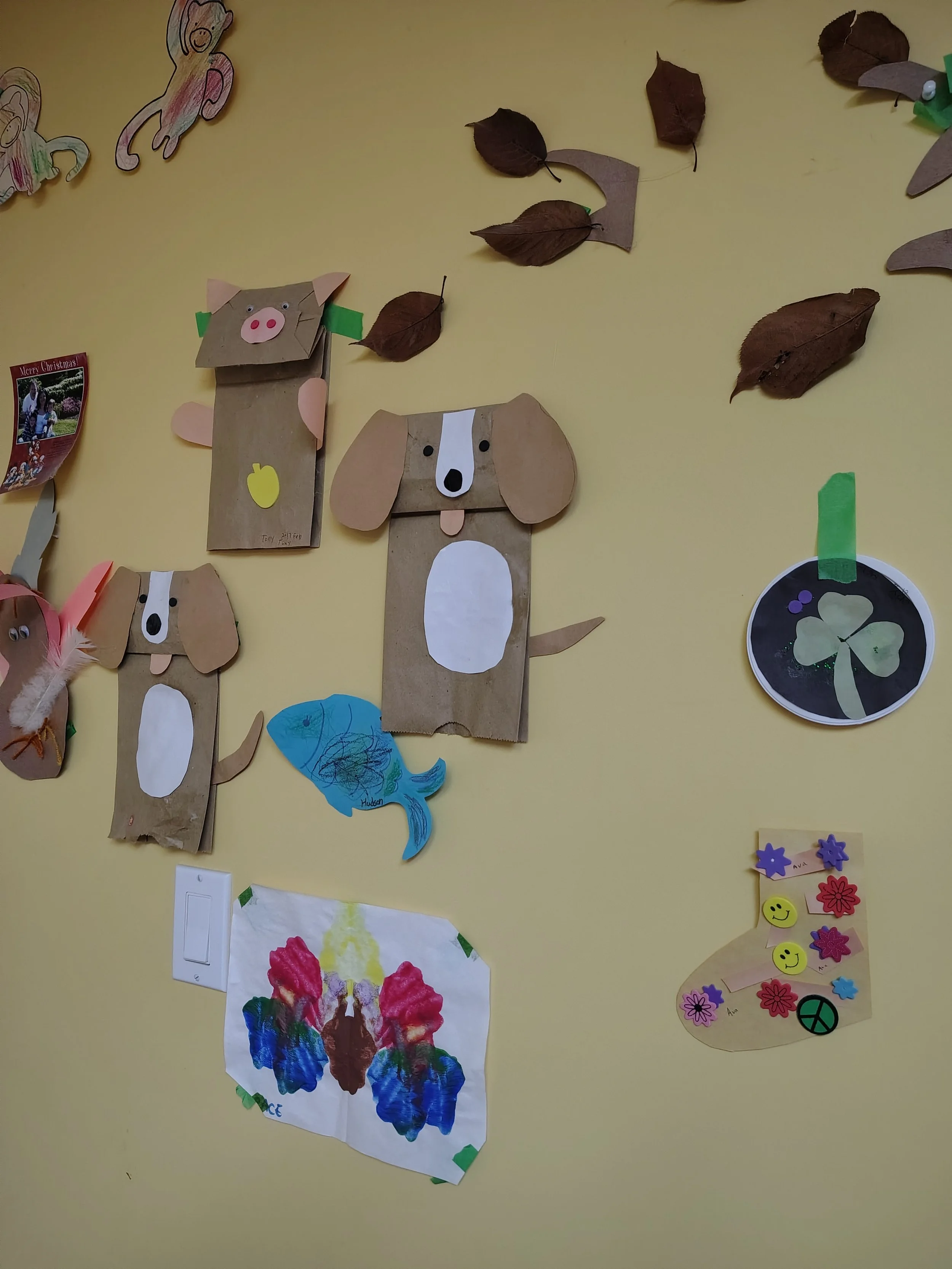 Children's artwork on a yellow wall, including paper crafts of dogs and a pig made from paper and tissue, a blue fish drawing, and a colorful paint drawing of a butterfly.