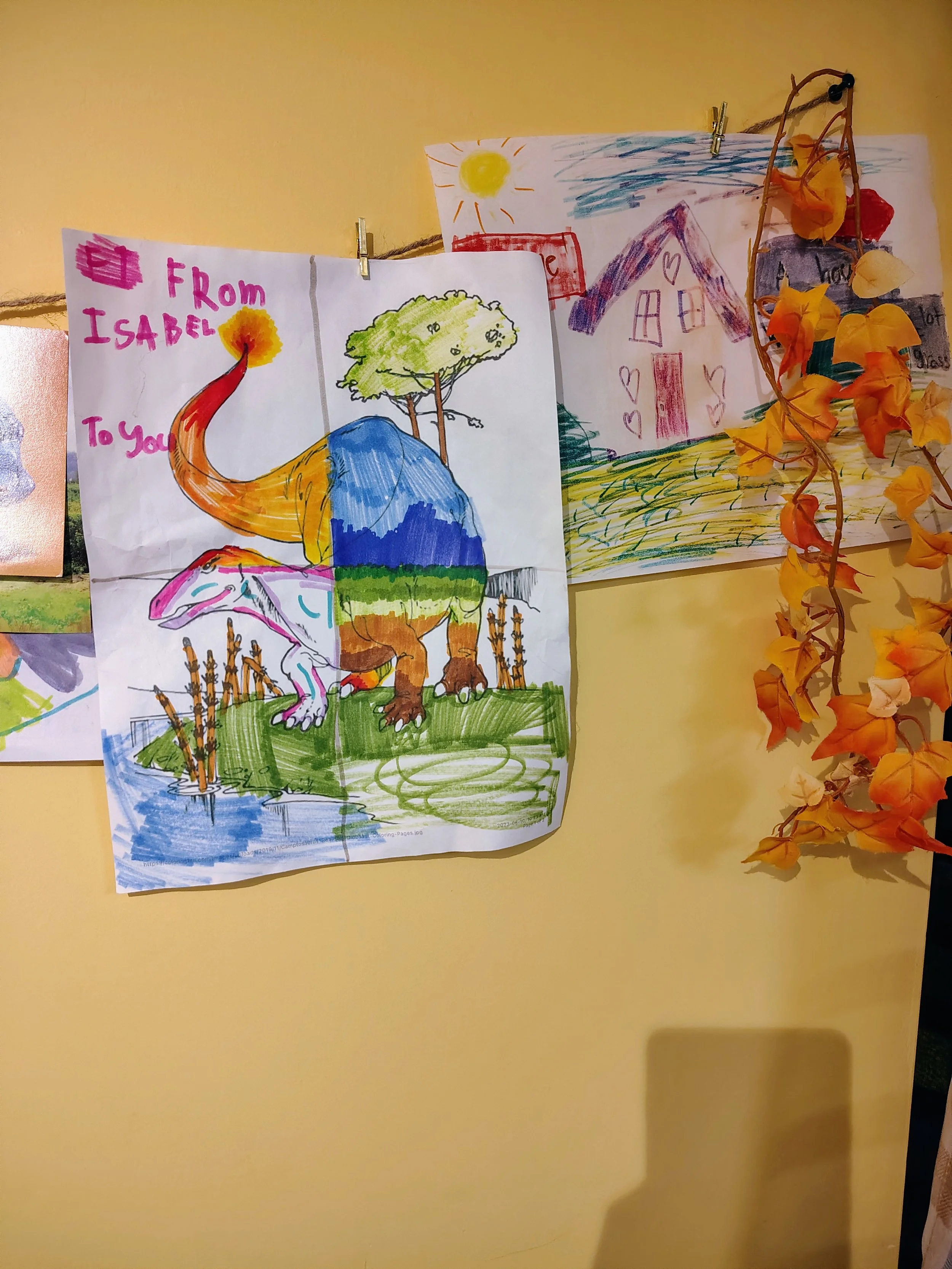 Colorful children's drawing of a dinosaur with trees, water, and the words 'From Isabel to You' written at the top; in the background, there are other children's drawings and some orange flowers.