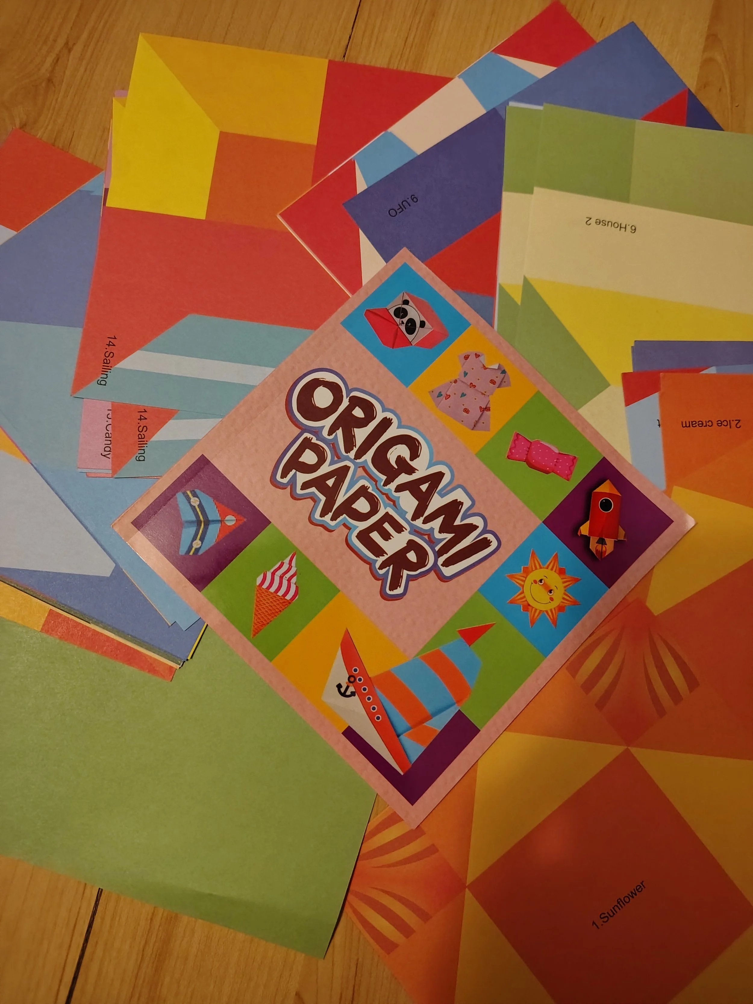Colorful origami paper sheets with various patterns and colors, and a pink booklet labeled "Origami Paper" featuring images of paper animals, a boat, and a sun.