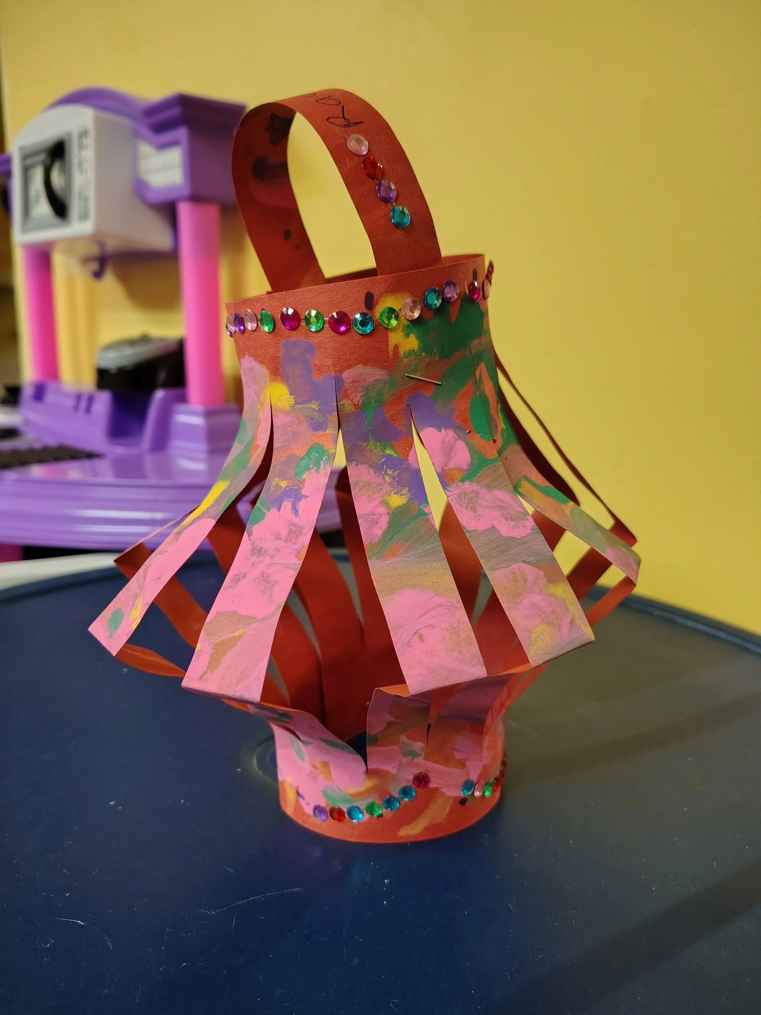 Colorful paper lantern decorated with rhinestones and paint, on a dark surface with a yellow wall and purple shelving in the background.