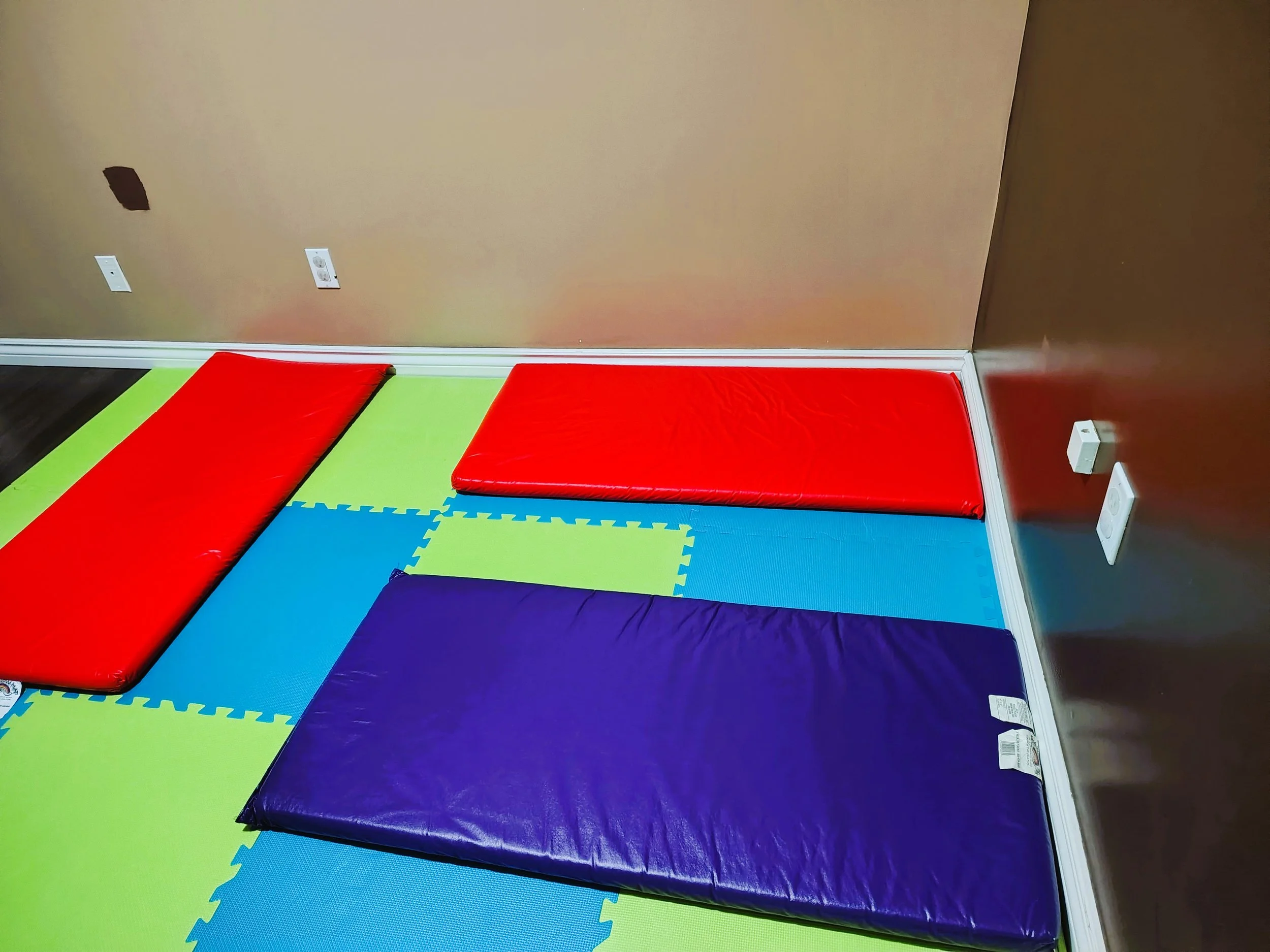 Colorful foam mats and red padding on the floor in a room with electrical outlets on the wall.