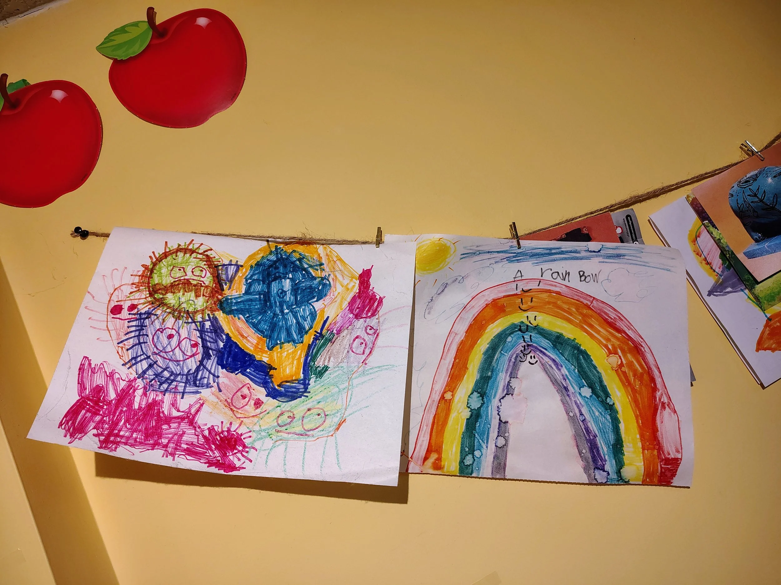 Colorful children's drawings on white paper attached to a string with small clips, one depicting a blue cloud and sun, and the other a rainbow with smiley faces, titled 'A rain bow'.