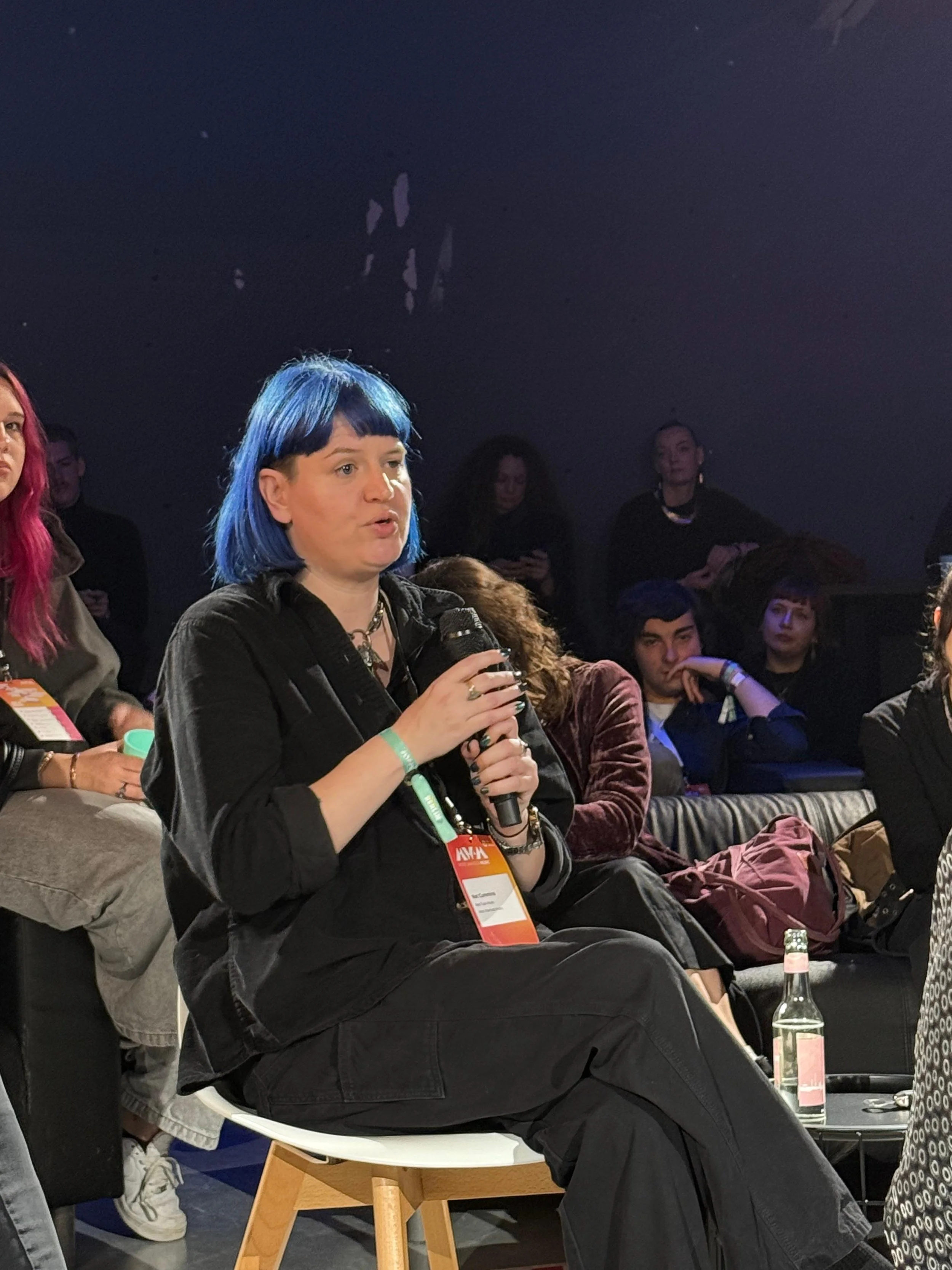 A woman with blue hair, wearing black clothing and a conference badge, holding a microphone and speaking at an indoor event.