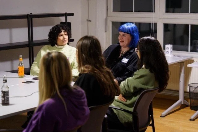 Five women sitting around a table in a room with large windows, a black metal rack, and a wooden floor. One woman with blue hair is smiling while talking to the others.