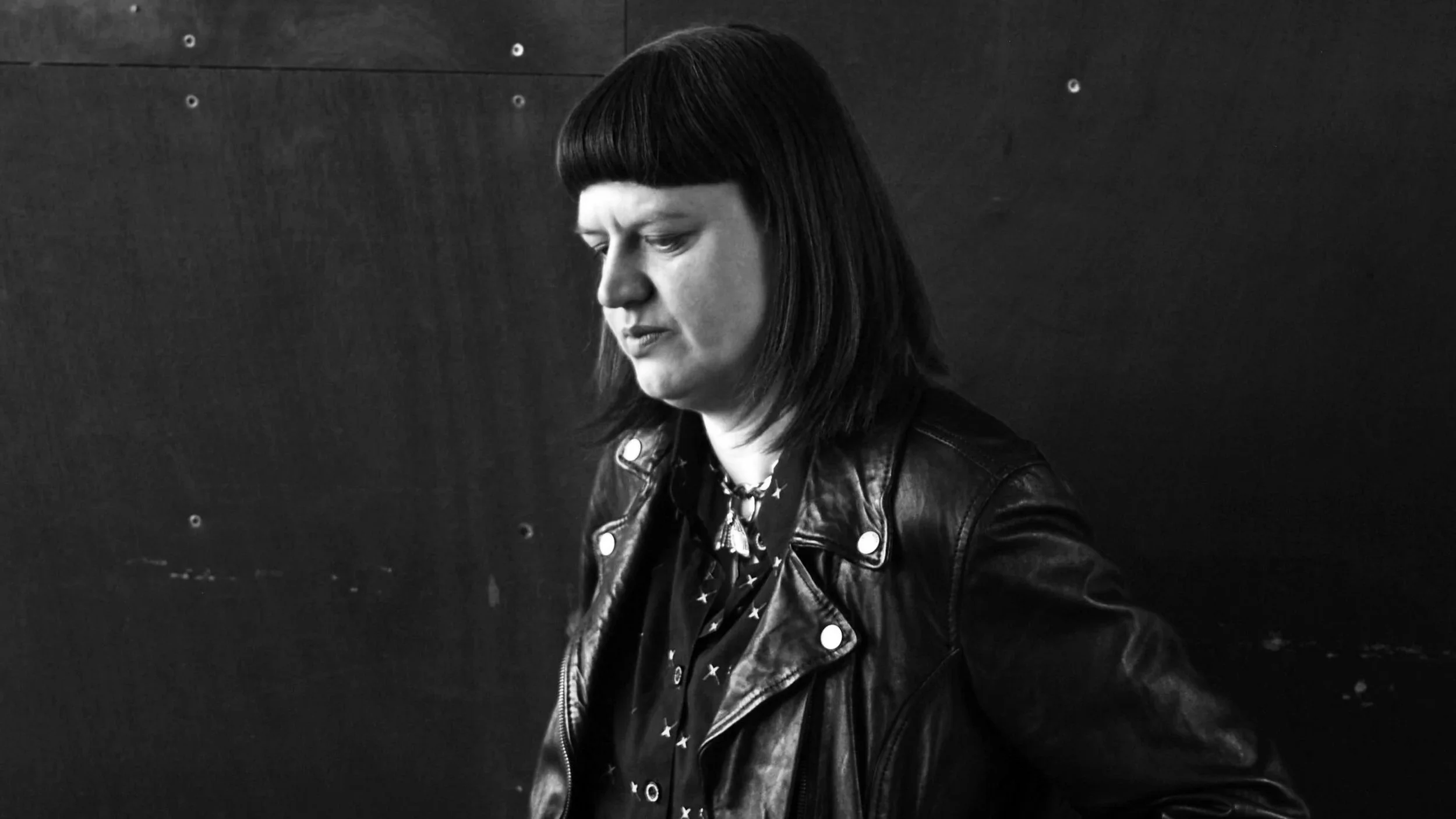 Black and white photograph of a woman with shoulder-length dark hair, wearing a leather jacket and a patterned shirt, standing against a dark background.