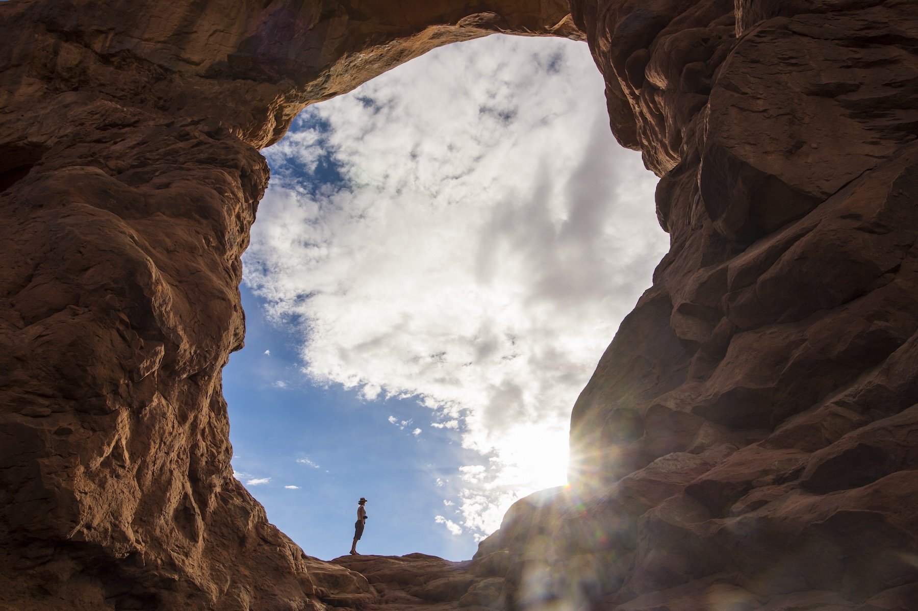 A person standing inside a large rocky canyon or crevice, looking towards the open sky with clouds, with sunlight shining through the rocks.