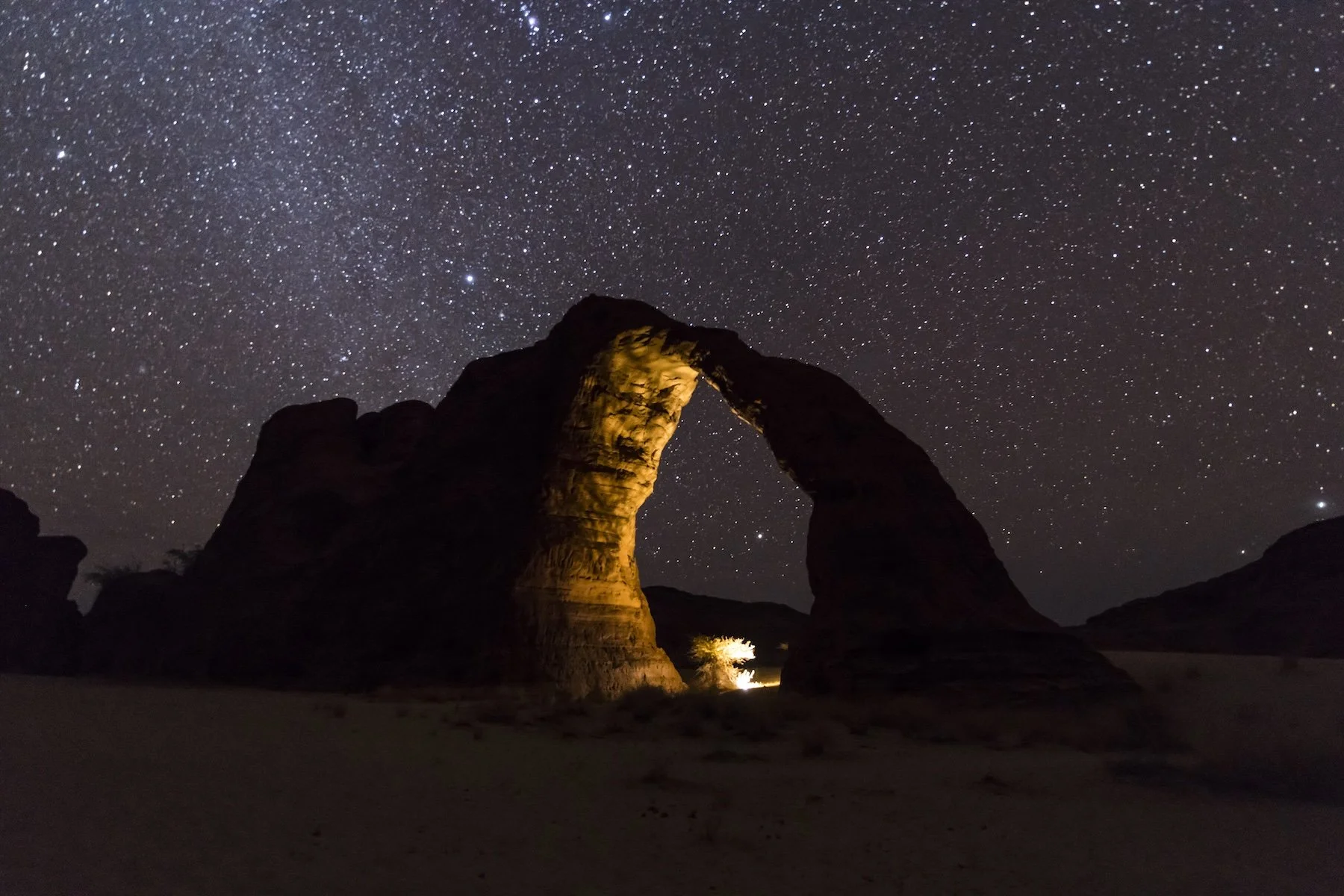 Night sky filled with stars over a rock formation with an arch illuminated by warm lights.