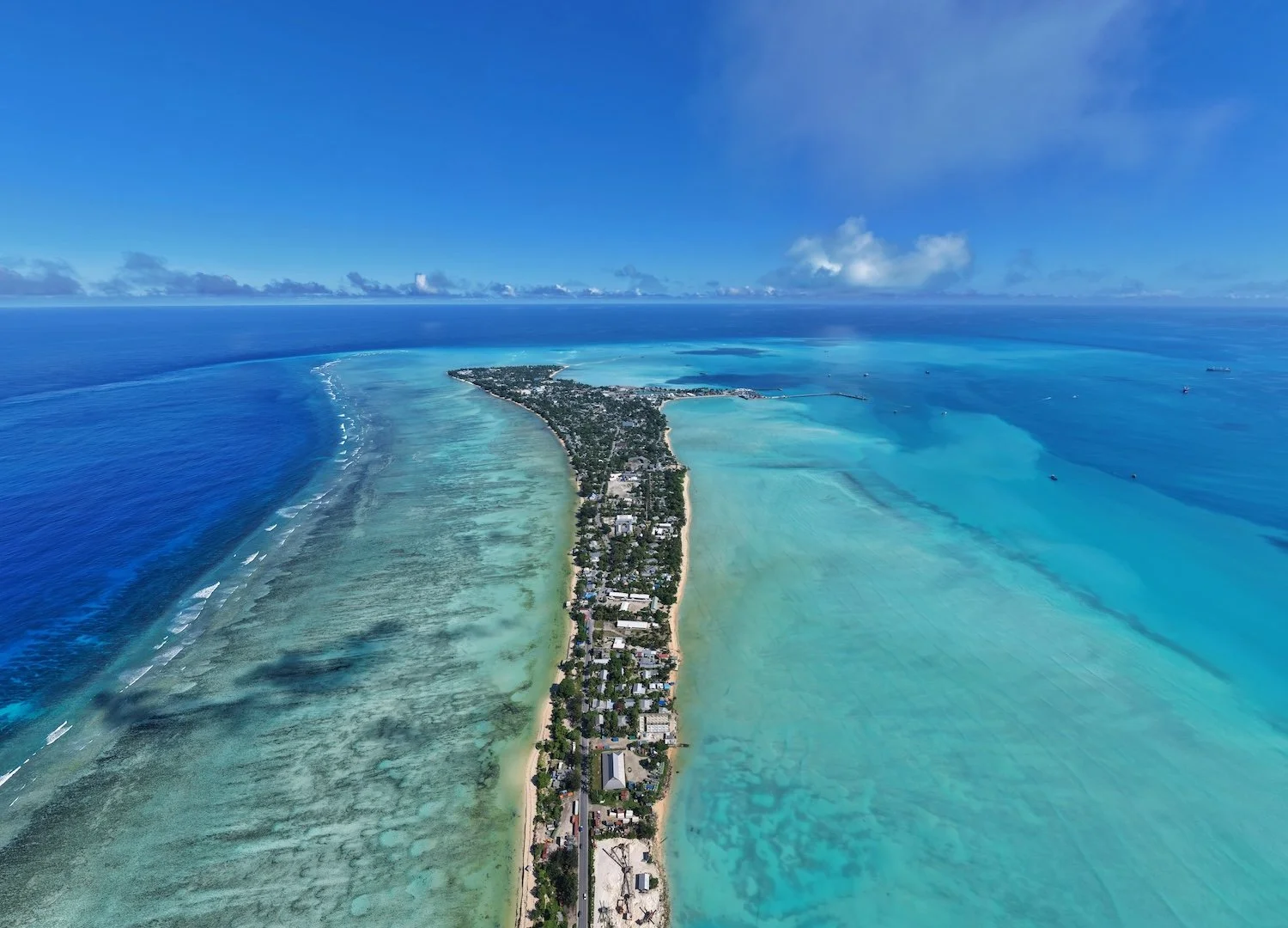 Aerial view of the capital of the Pacific island nation of Kiribati, Tarawa, with buildings and houses along a long, narrow atoll, surrounded by pristine blues of the Pacific Ocean.