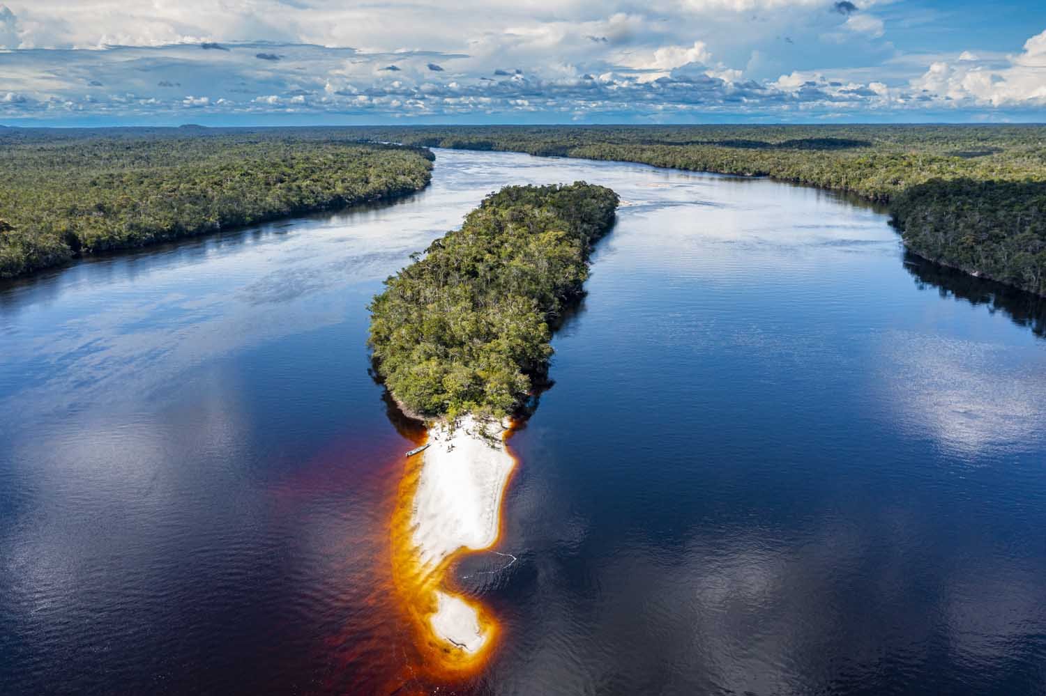 Aerial view of a river with orange stains on the shoreline and an island covered in trees in the middle of the waterway, surrounded by lush green forest and cloudy sky.