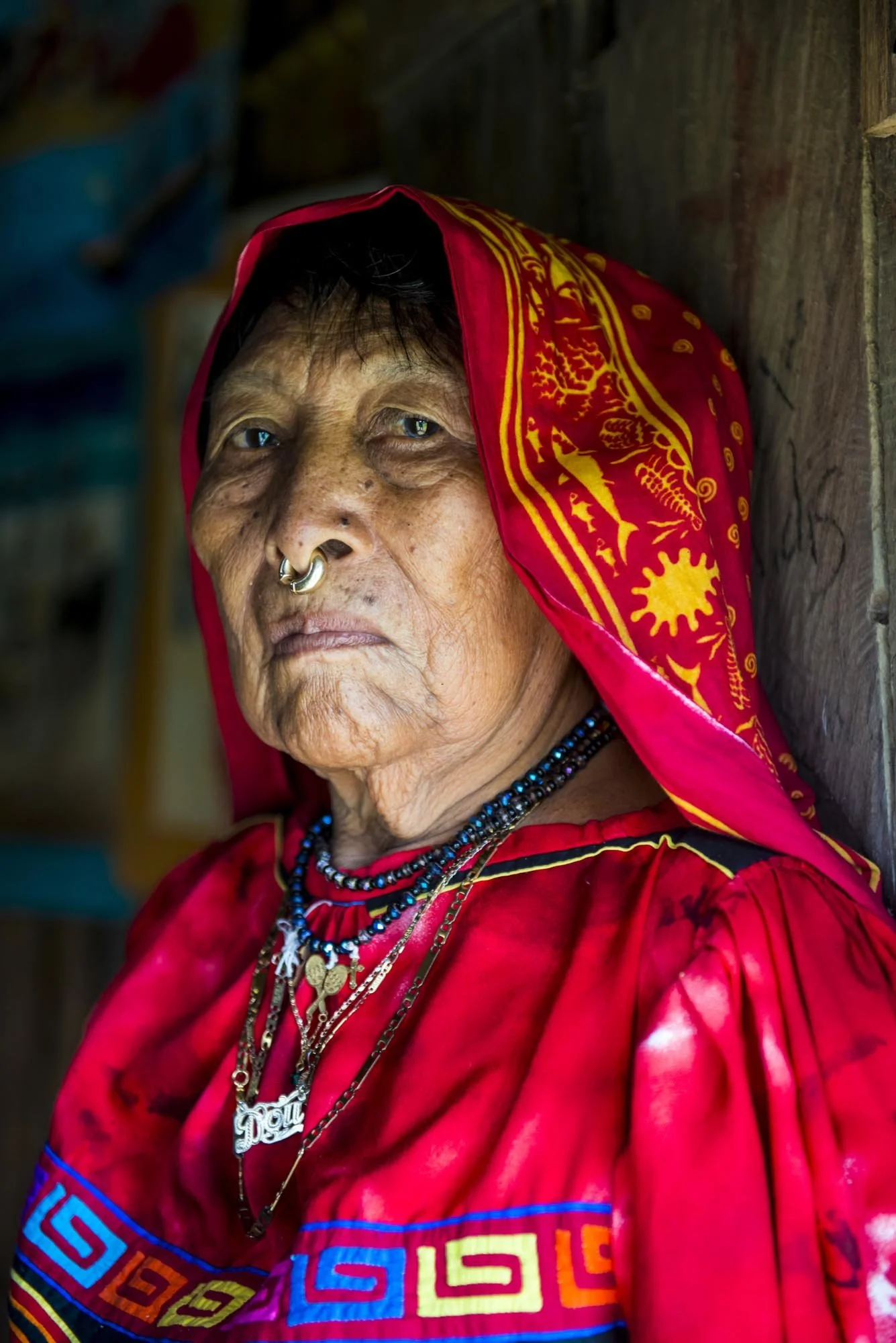An elderly woman wearing traditional red clothing and a headscarf decorated with yellow patterns, adorned with multiple necklaces and a nose ring, sitting against a wooden background.
