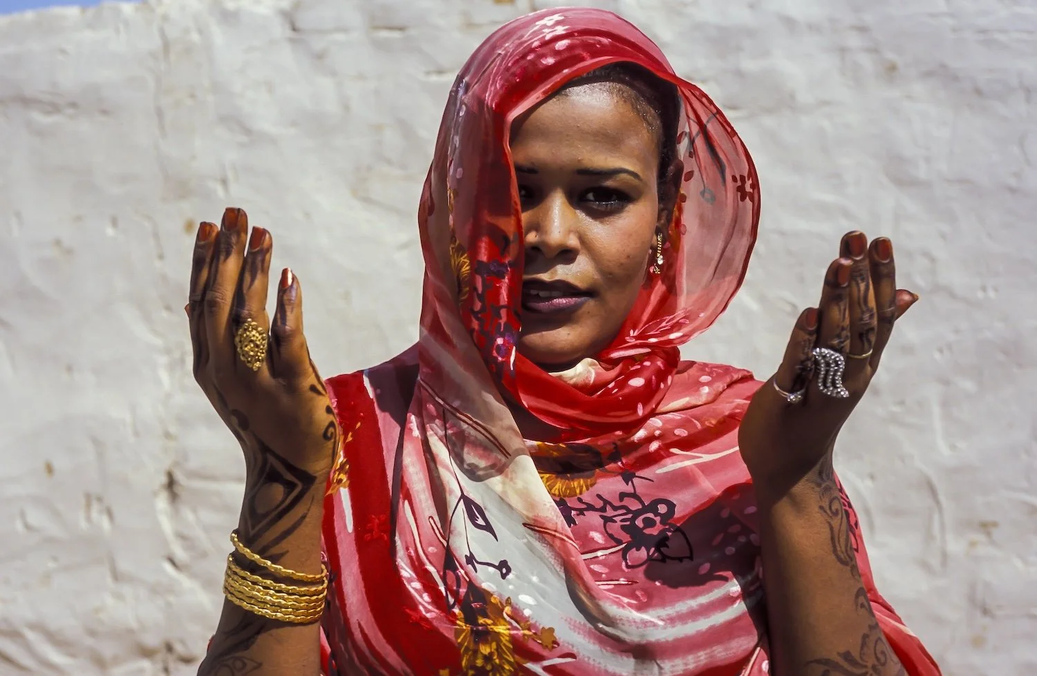 Woman wearing a colorful red and pink headscarf, with tattoos on her arms, earrings, and jewelry, standing against a white wall, with her hands raised in a gesture.