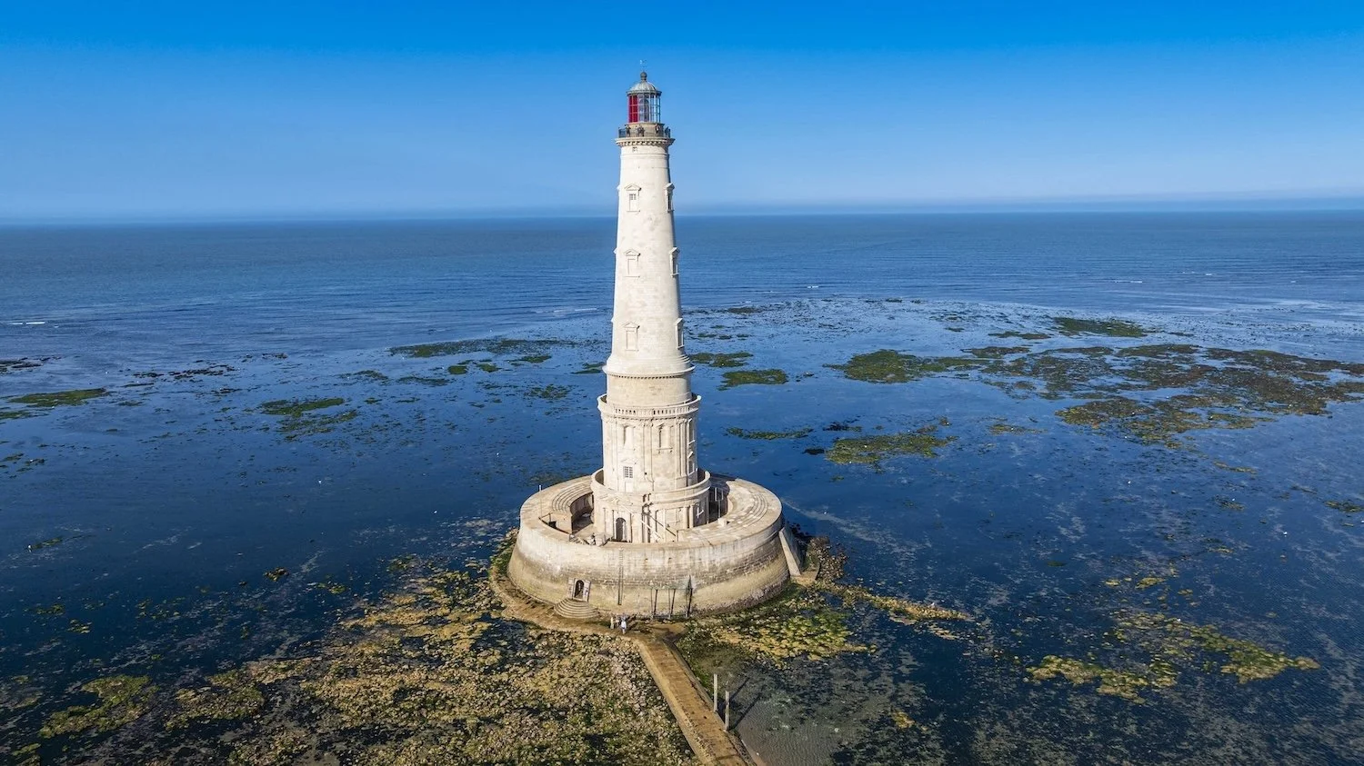 A tall white lighthouse on a small rocky island surrounded by water with patches of seaweed, under a clear blue sky.