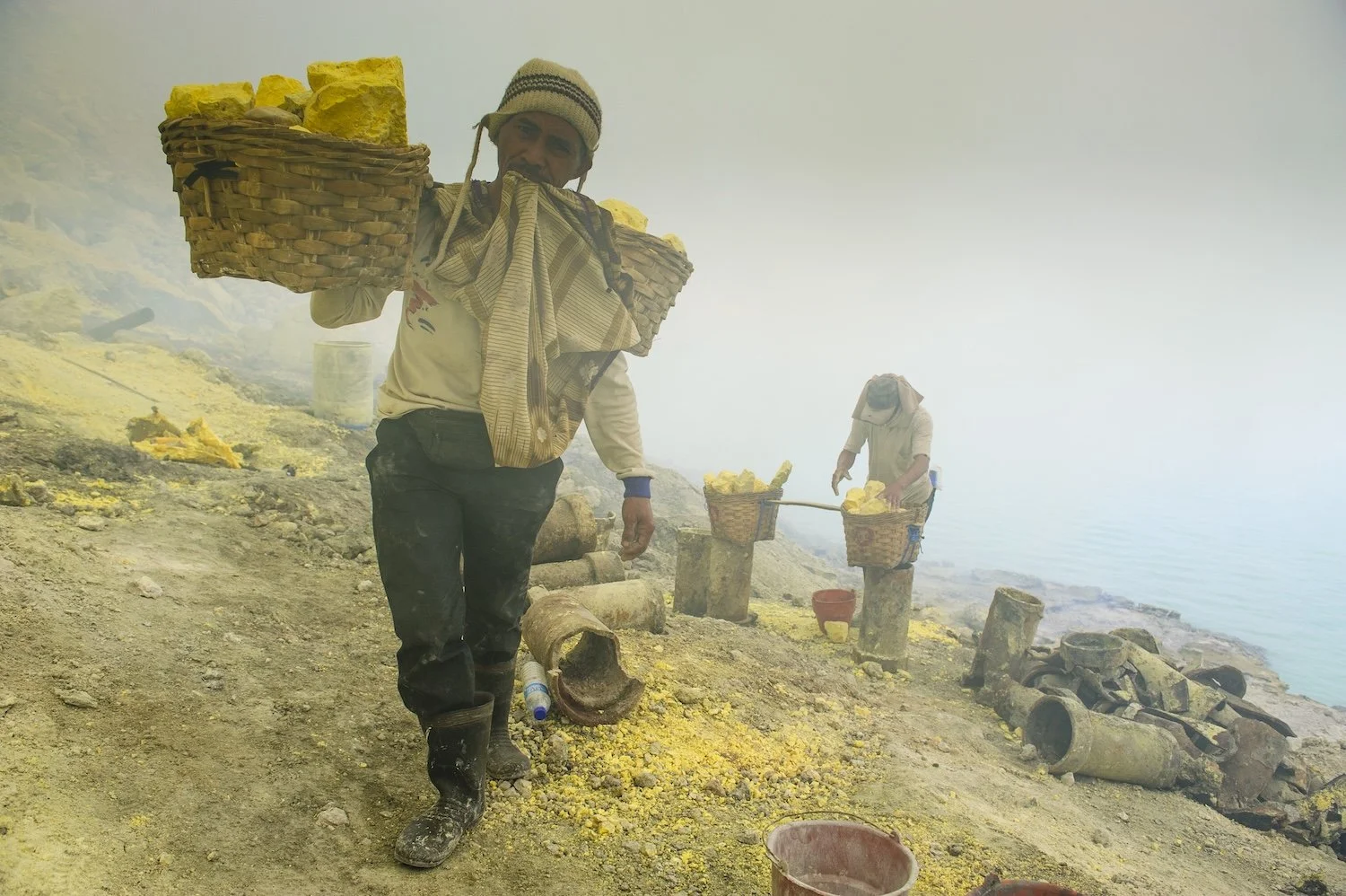 Two men are harvesting sulfur on volcanic terrain near a body of water. They are carrying baskets filled with yellow sulfur.