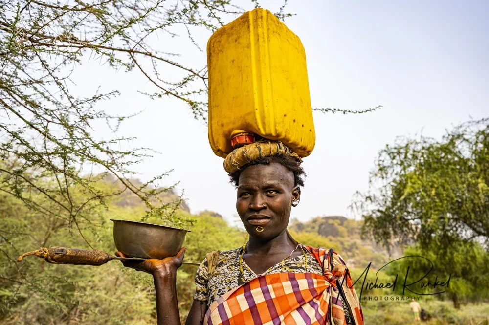 A portrait of a woman of the Larim tribe in South Sudan, Africa, colorfully dressed and carrying a canister of water on her head.