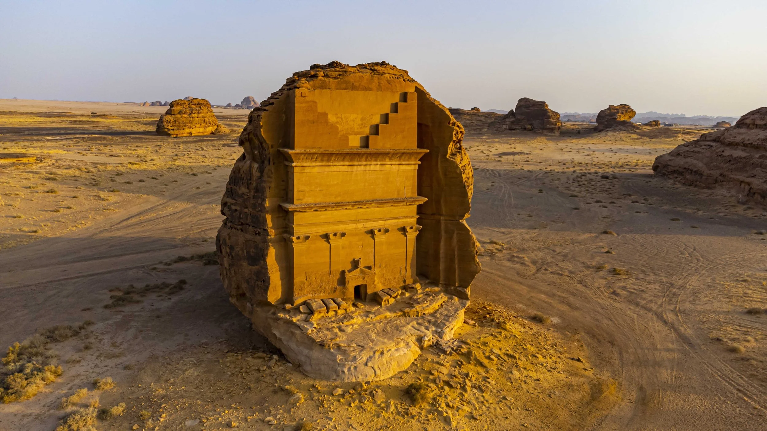 Sandstone monument resembling a house with stairs on a desert landscape at sunset, surrounded by scattered rock formations.