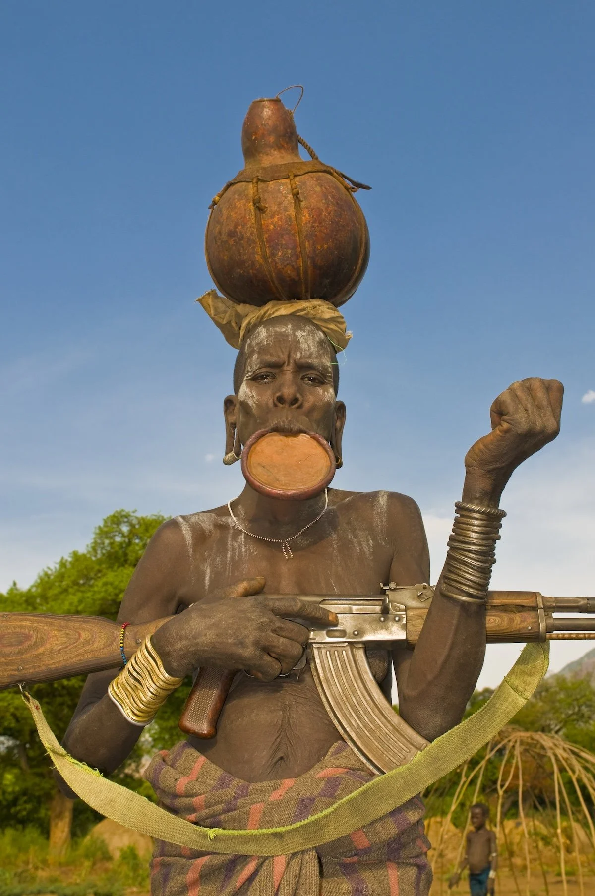 A Mursi man with a traditional lip plate, a gourd hat, decorative body art and holding a rifle, lower Omo Valley in southeastern Ethiopia, Africa.