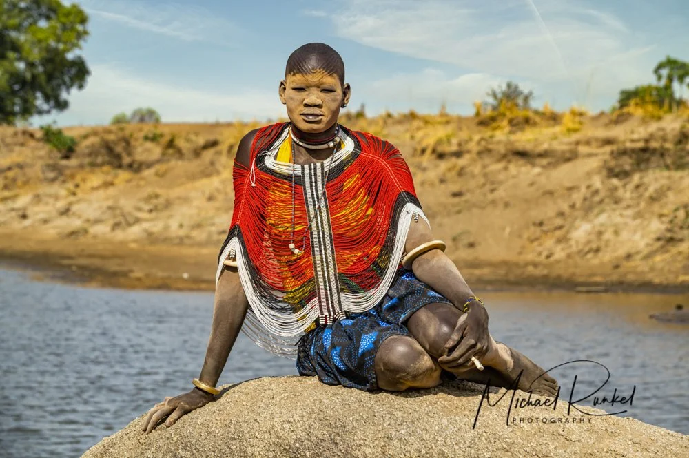 A young person from an indigenous African tribe, wearing traditional beadwork and colorful clothing, sits on a rock by a river with a landscape of dry hills and trees in the background.