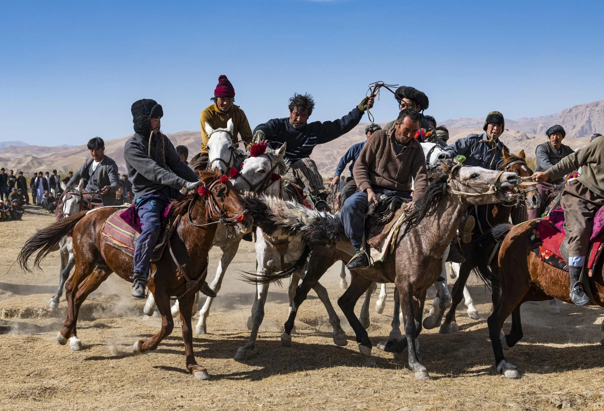 Group of people riding horses across a desert landscape, involved in a gathering or celebration, with mountains in the background under a clear blue sky.