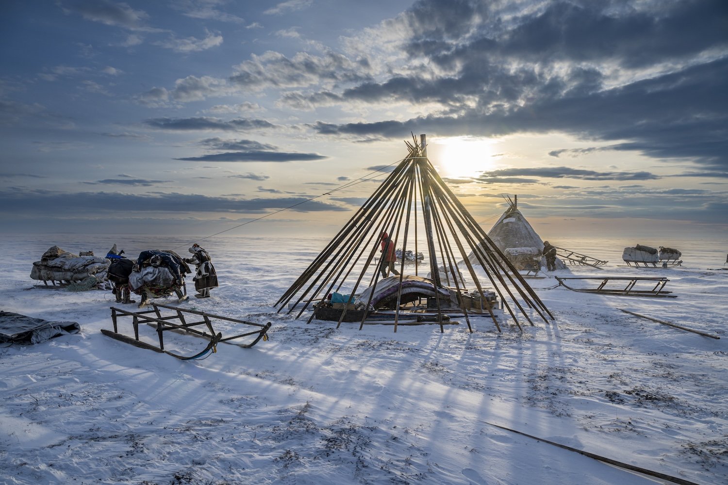 A snowy landscape with several teepee-style structures made of wooden poles, some covered with animal skins or fabric, set up on a frozen surface. People are working around the structures, and sleds loaded with supplies are visible in the scene. The 
