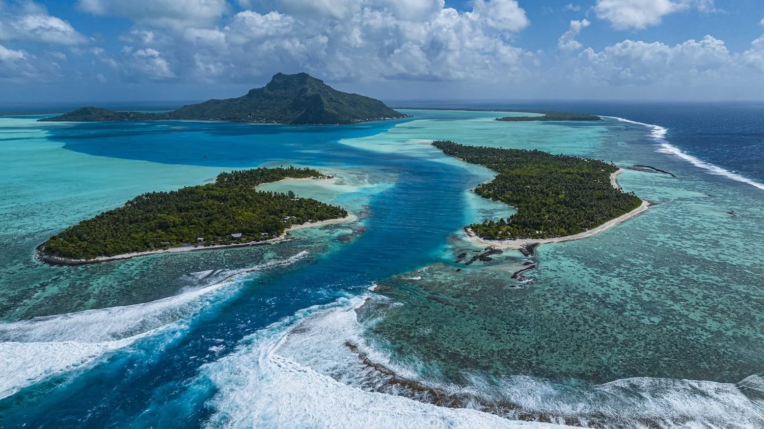 An aerial view of a tropical island surrounded by vibrant blue waters and coral reefs, with a mountain in the background under a partly cloudy sky.