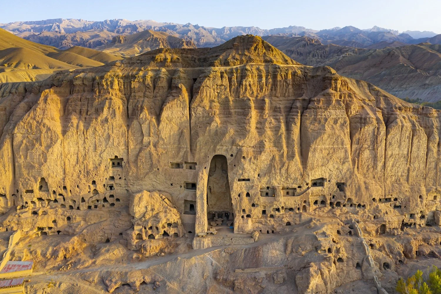 Ancient cave dwellings carved into a large sandstone cliff in a desert landscape, with mountains in the background.
