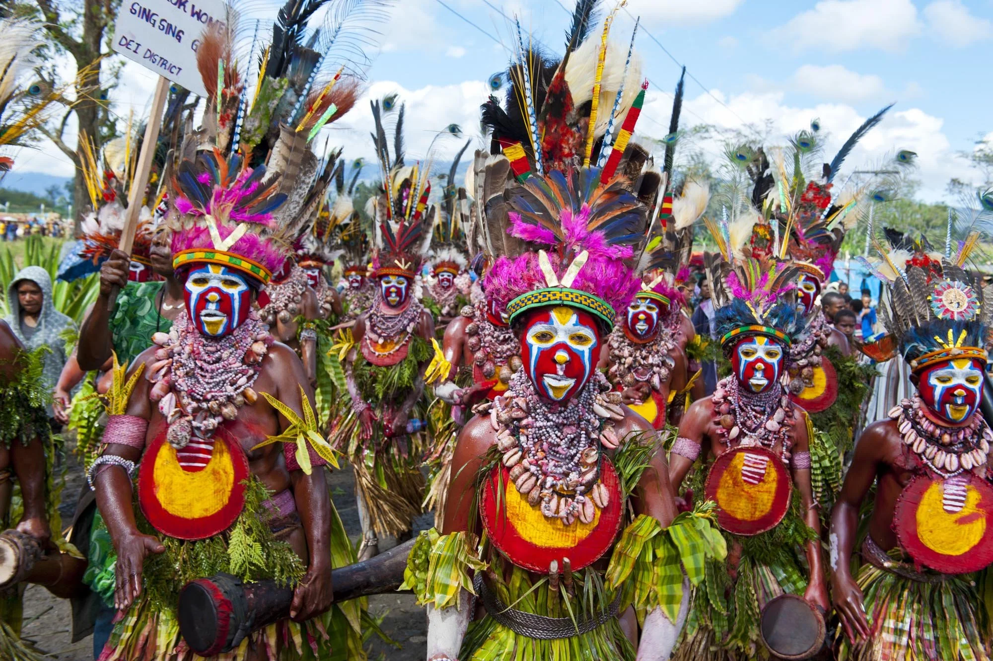 People in traditional tribal costumes, wearing face paint, feathers, jewelry, and colorful clothing, participating in a cultural event or parade outdoors on a sunny day.