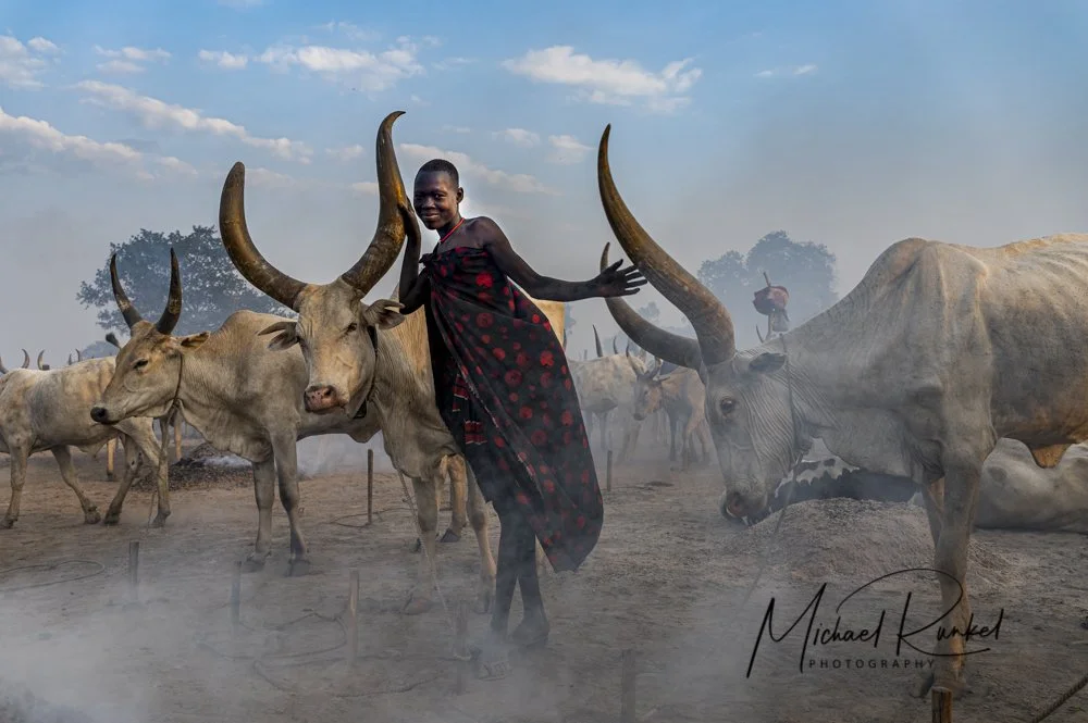A person in traditional clothing standing among a herd of large, horned cattle outdoors during the daytime with a partly cloudy sky.