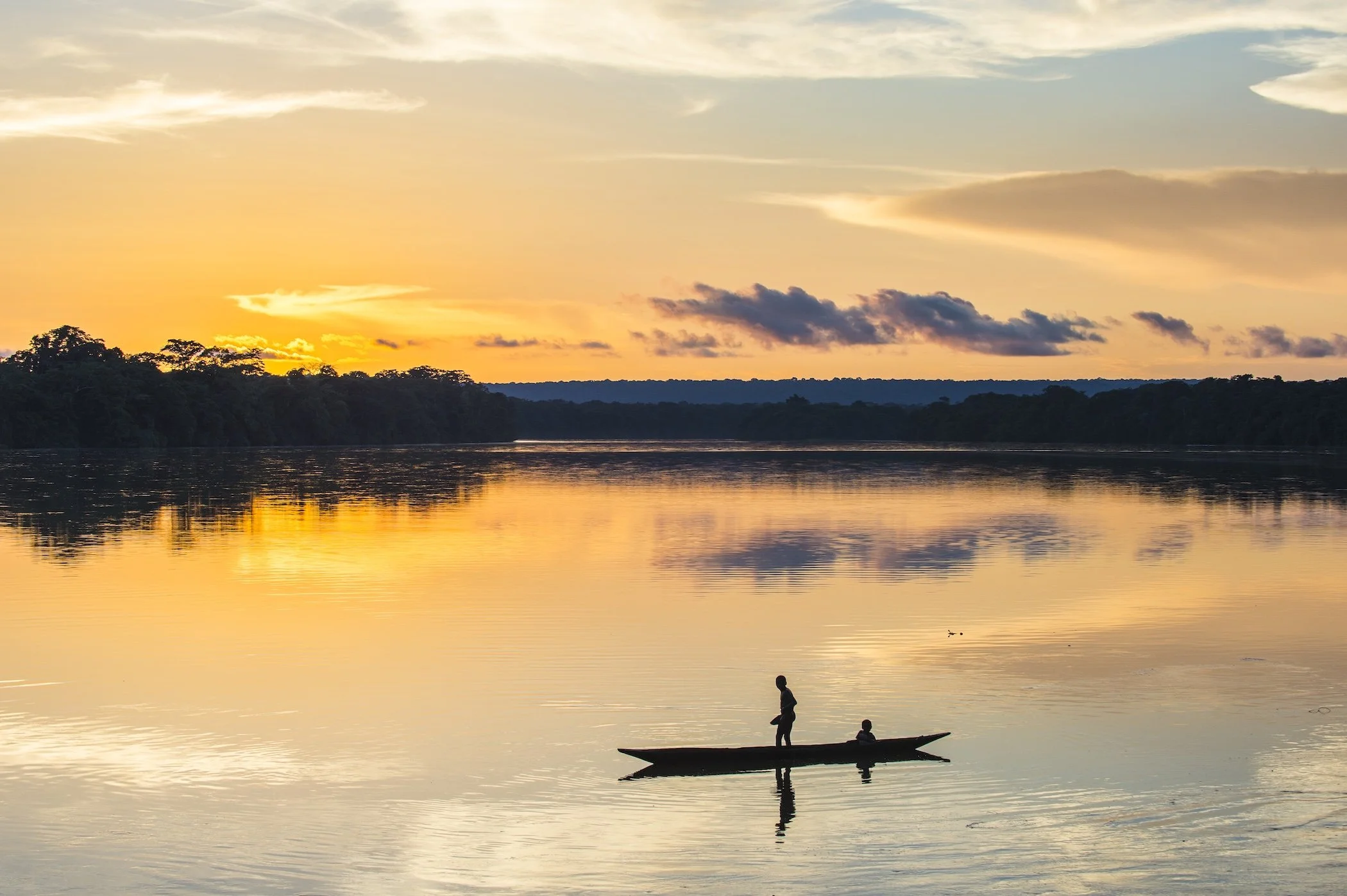 A calm river at sunset with four people in a small boat, two adults and two children, and a dramatic sky with clouds and a golden glow.