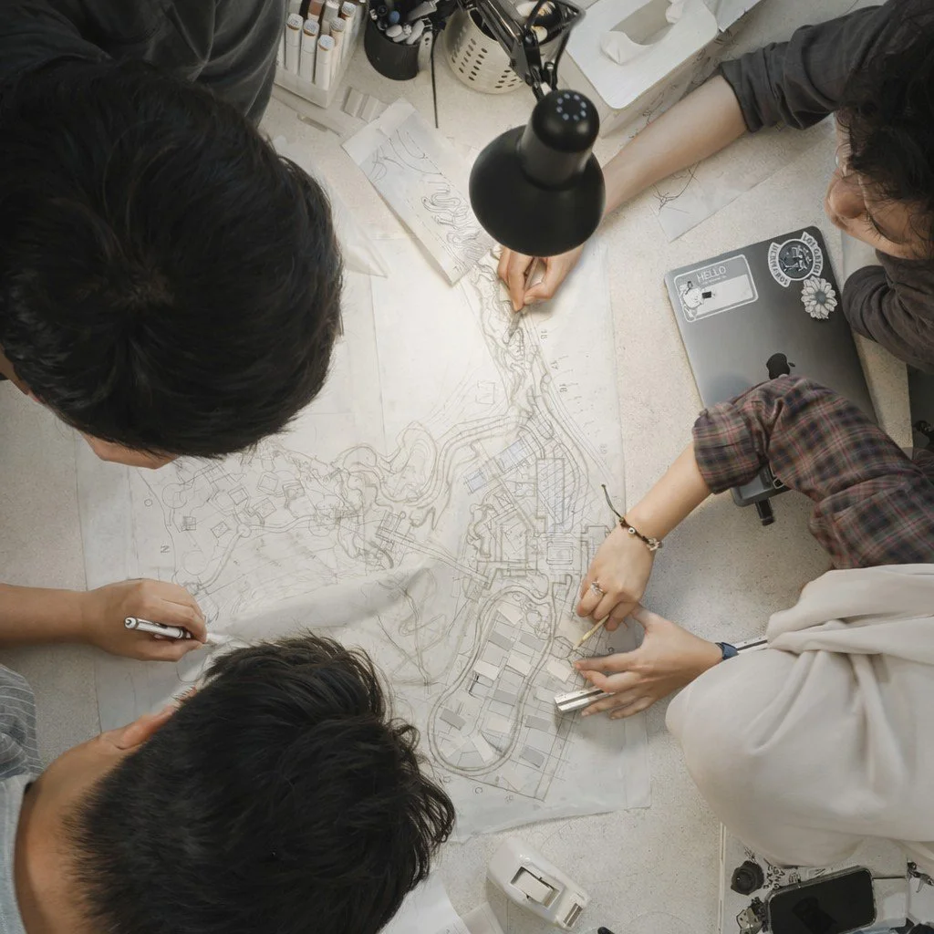 Group of people working on a large blueprint of an urban development plan on a table, with drafting tools, laptop, and drawing materials around them.