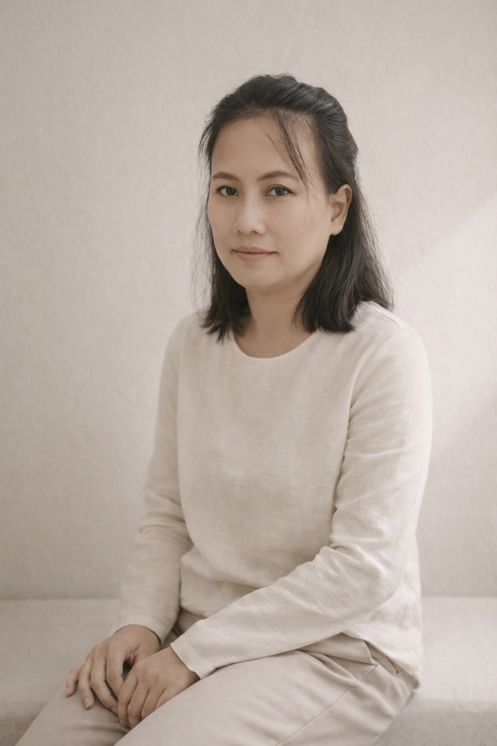 A woman sitting on a beige couch with her hands folded on her lap, wearing a light-colored long-sleeve shirt, with dark hair and a gentle smile, against a plain beige wall.